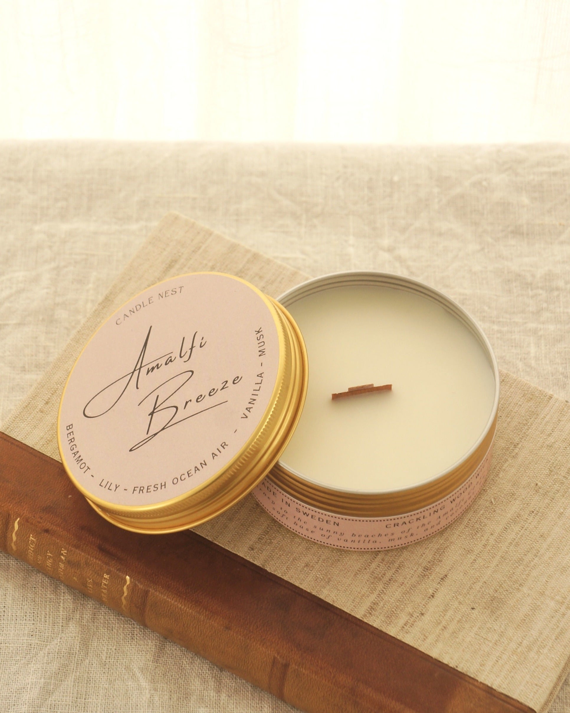 Ett doftljus i en rund guldfärgad burk av metall står på en bok. Boken är av linnetyg med en rygg i läder. Ljusets lock står lutad mot ljuset och på det finns ljusets namn skrivet med snirklig text "Amalfi Breeze". Mitt i ljuset syns en veke av trä. Bilden är tagen snett uppifrån.