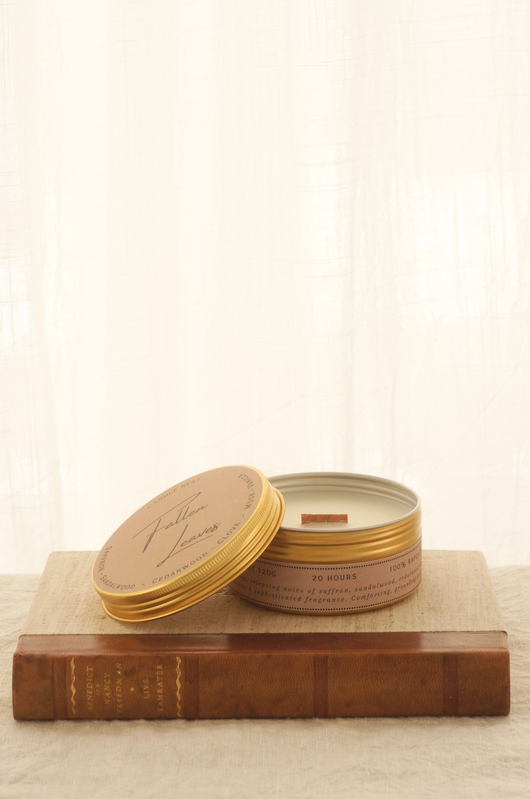 Ett doftljus i en rund guldfärgad burk av metall står på en bok. Boken är av linnetyg med en rygg i läder. Ljusets lock står lutad på ljuset på på det finns ljusets namn skrivet med snirklig text "Fallen Leaves". Mitt i ljuset syns en veke av trä. Bild tagen från sidan.