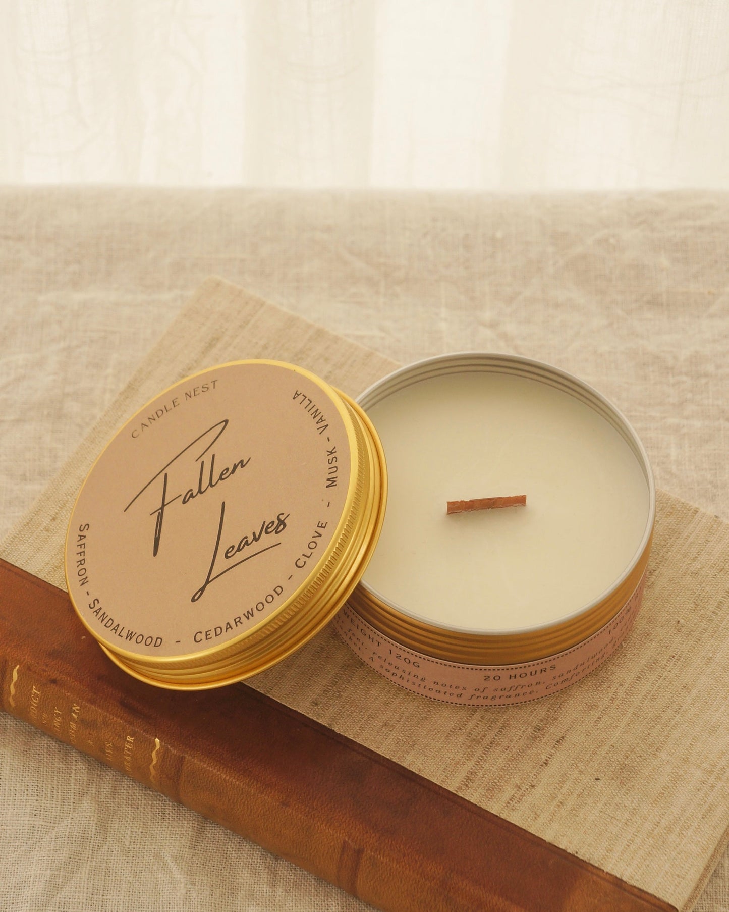 Ett doftljus i en rund guldfärgad burk av metall står på en bok. Boken är av linnetyg med en rygg i läder. Ljusets lock står lutad på ljuset på på det finns ljusets namn skrivet med snirklig text "Fallen Leaves". Mitt i ljuset syns en veke av trä. 