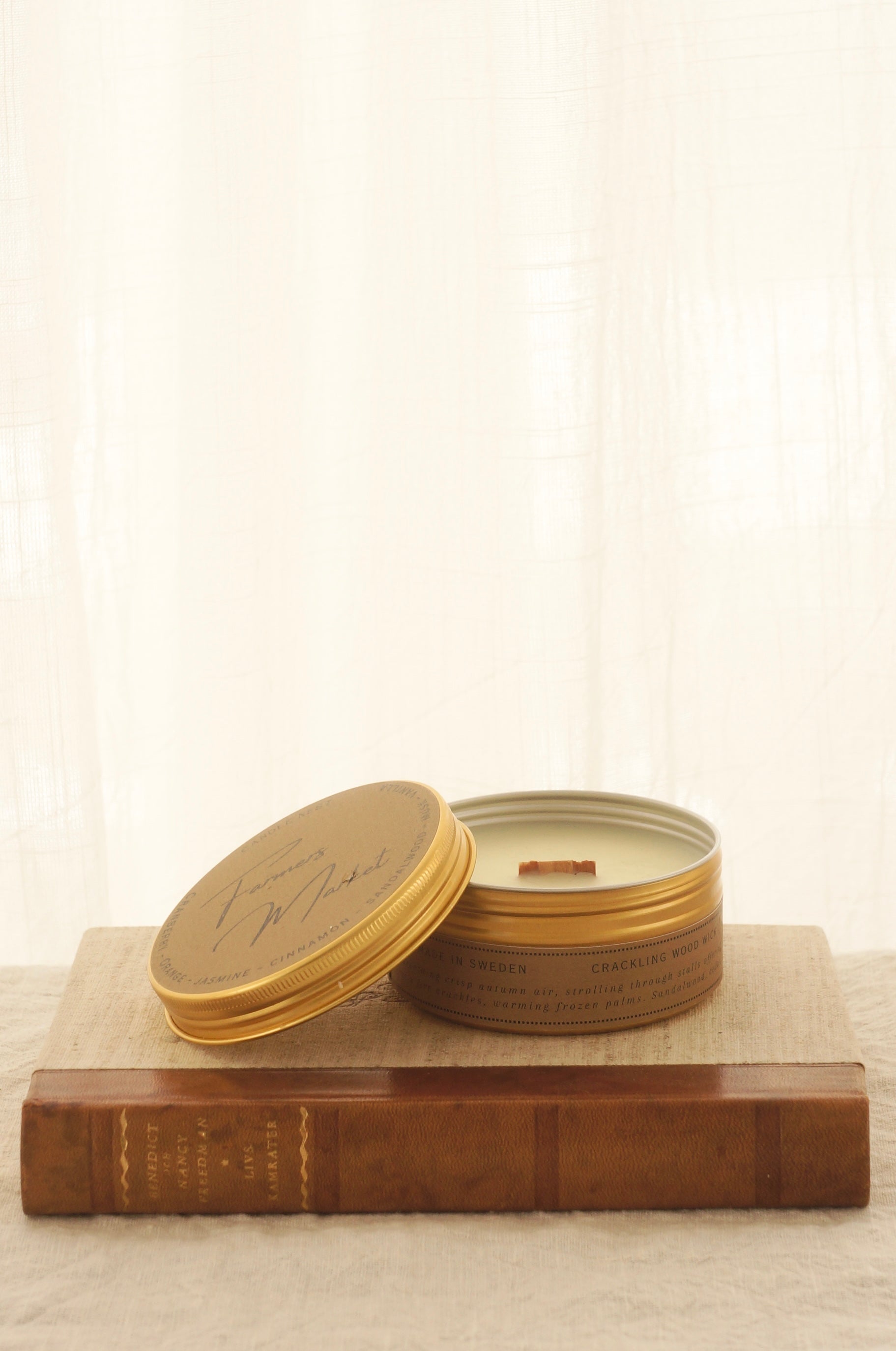 Ett doftljus i en rund guldfärgad burk av metall står på en bok. Boken har pärmar av av linnetyg med en rygg i läder. Ljusets lock står lutad på ljuset på på det finns ljusets namn skrivet med snirklig text "Farmers Market". Mitt i ljuset syns en veke av trä. Boken ligger på en duk av linnetyg. Bilden är tagen från sidan.