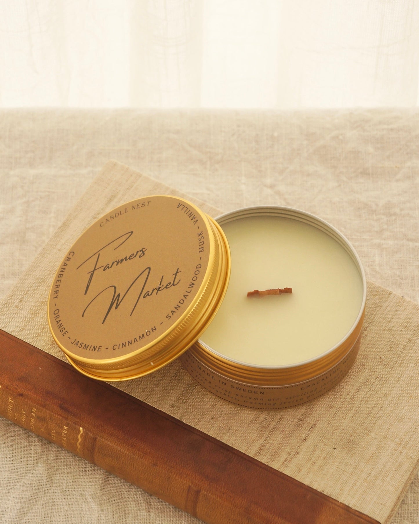 Ett doftljus i en rund guldfärgad burk av metall står på en bok. Boken har pärmar av av linnetyg med en rygg i läder. Ljusets lock står lutad på ljuset på på det finns ljusets namn skrivet med snirklig text "Farmers Market". Mitt i ljuset syns en veke av trä. Boken ligger på en duk av linnetyg. Bilden är tagen snett uppifrån.