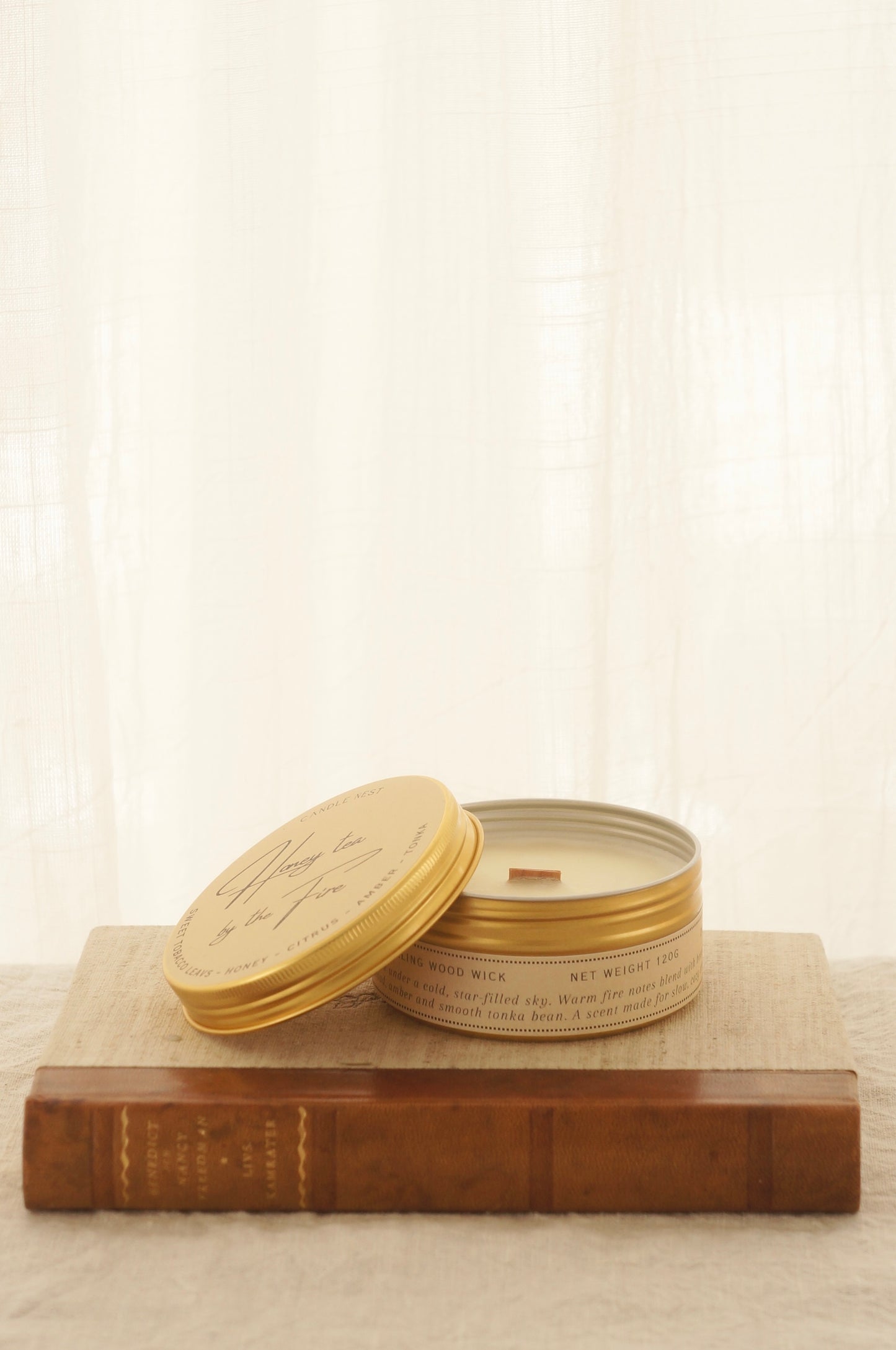 Ett doftljus i en rund guldfärgad burk av metall står på en bok. Boken är av linnetyg med en rygg i läder. Ljusets lock står lutad på ljuset på på det finns ljusets namn "Honey tea at the Fire" skrivet med snirklig text. Boken står på en beige duk av linnetyg. Mitt i ljuset syns en bed veke av trä. Bilden är tagen från sidan. 