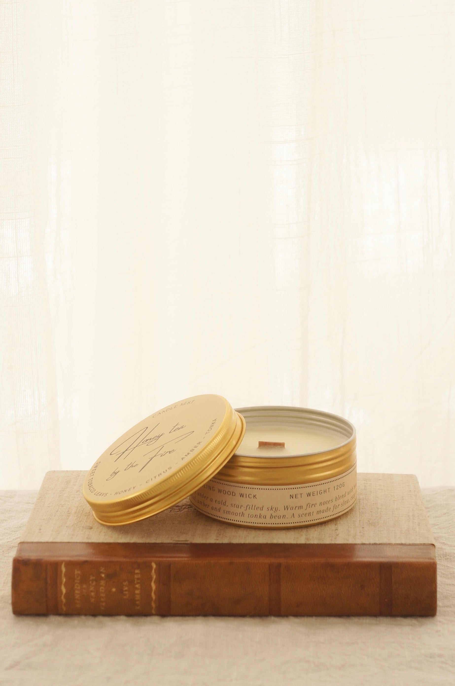 Ett doftljus i en rund guldfärgad burk av metall står på en bok. Boken är av linnetyg med en rygg i läder. Ljusets lock står lutad på ljuset på på det finns ljusets namn "Honey tea at the Fire" skrivet med snirklig text. Boken står på en beige duk av linnetyg. Mitt i ljuset syns en bed veke av trä. Bilden är tagen från sidan. 