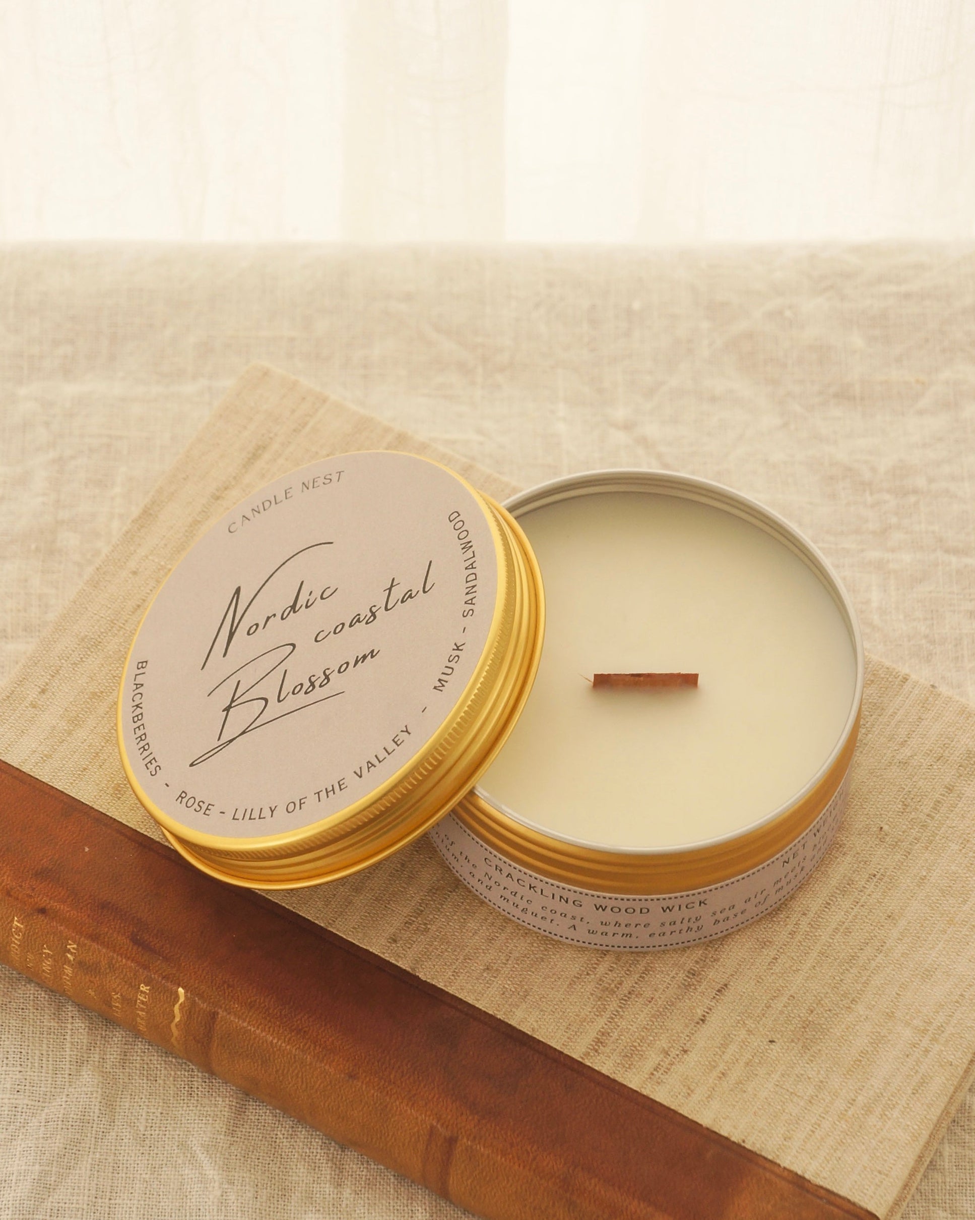 Ett doftljus i en rund guldfärgad burk av metall står på en bok. Boken är av linnetyg med en rygg i läder. Ljusets lock står lutad på ljuset på på det finns ljusets namn skrivet med snirklig text "Nordic coastal blossom". Mitt i ljuset syns en veke av trä.  Bilden är tagen snett uppiifrån.