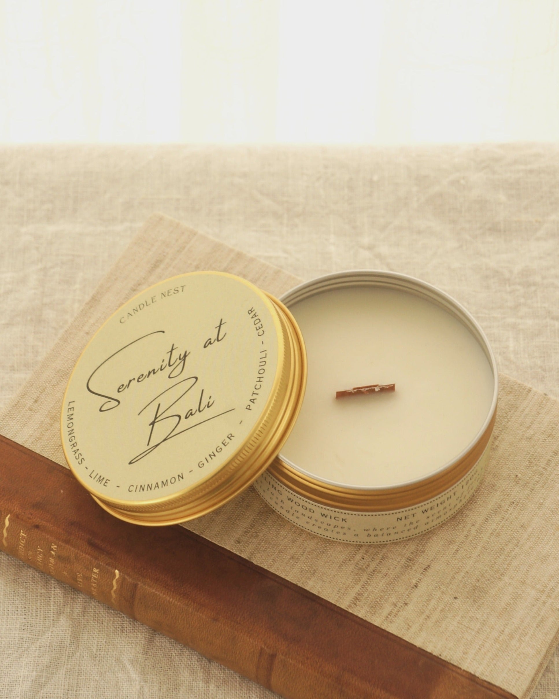 Ett doftljus i en rund guldfärgad burk av metall står på en bok. Boken är av linnetyg med en rygg i läder. Ljusets lock står lutad mot ljuset och på det finns ljusets namn skrivet med snirklig text "Serenity at Bali". Mitt i ljuset syns en veke av trä. Bilden är tagen snett uppifrån.