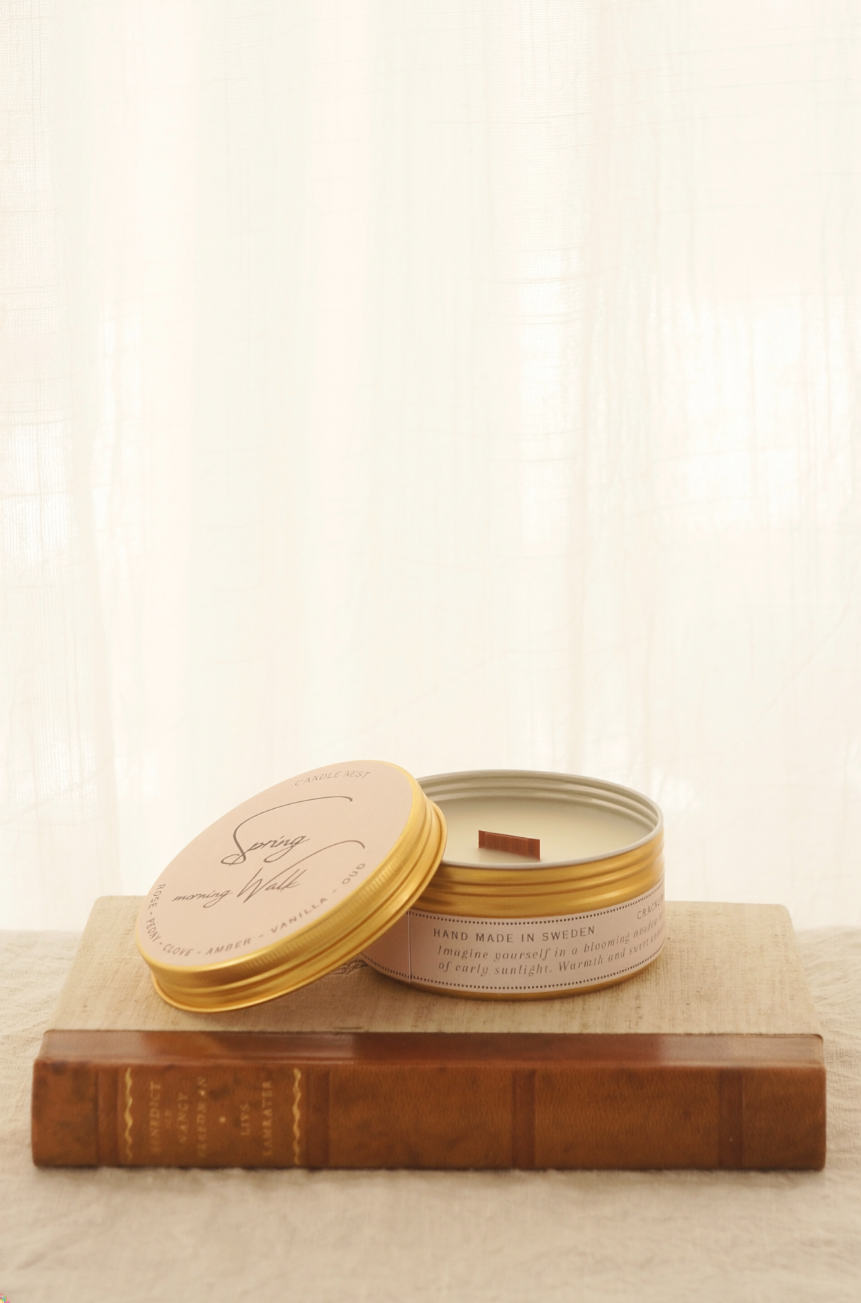 Ett doftljus i en rund guldfärgad burk av metall står på en bok. Boken är av linnetyg med en rygg i läder. Ljusets lock står lutad på ljuset på på det finns ljusets namn "Spring morning walks" skrivet med snirklig text. Boken står på en beige duk av linnetyg. Mitt i ljuset syns en bed veke av trä. Bilden är tagen från sidan.