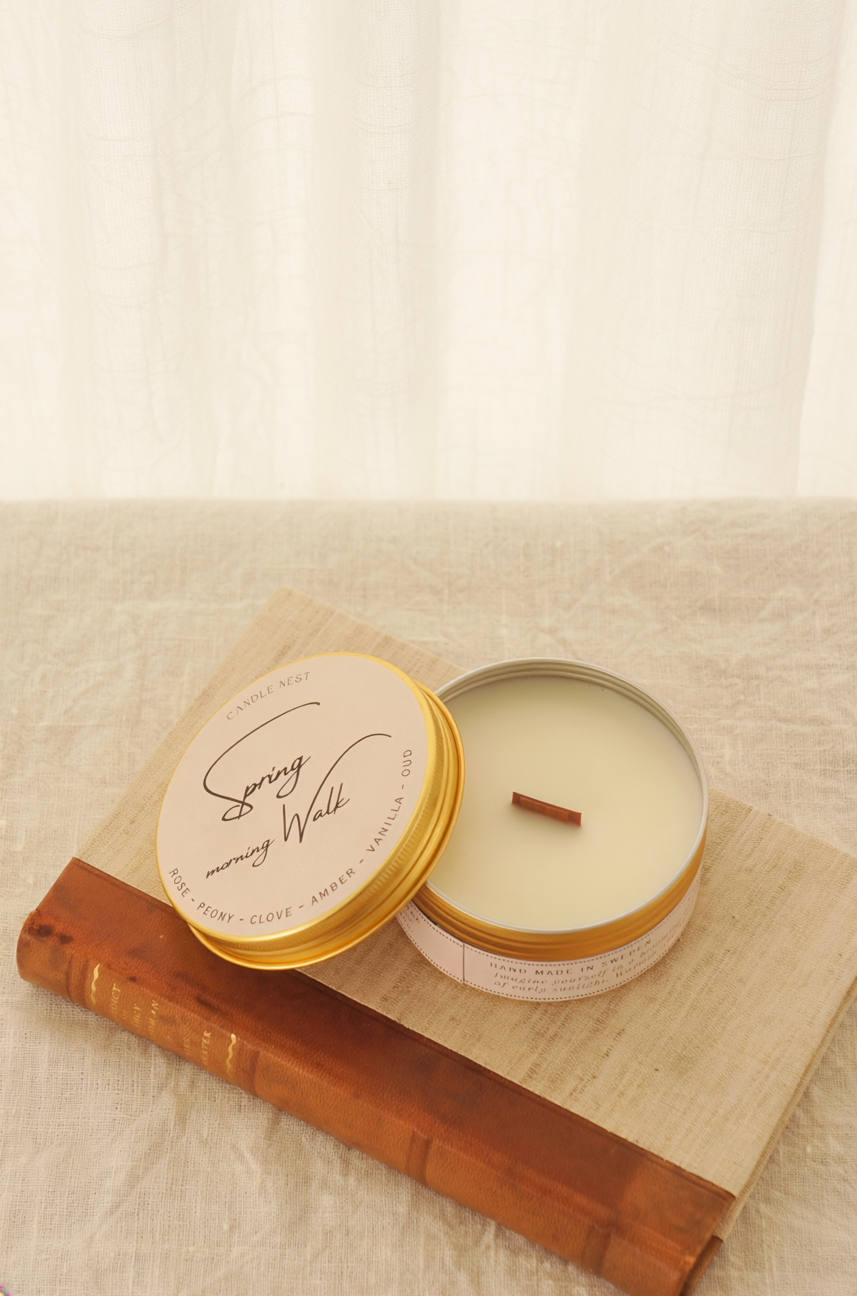Ett doftljus i en rund guldfärgad burk av metall står på en bok. Boken är av linnetyg med en rygg i läder. Ljusets lock står lutad på ljuset på på det finns ljusets namn "Spring morning walks" skrivet med snirklig text. Boken står på en beige duk av linnetyg. Mitt i ljuset syns en bed veke av trä. Bilden är tagen snett uppifrån.