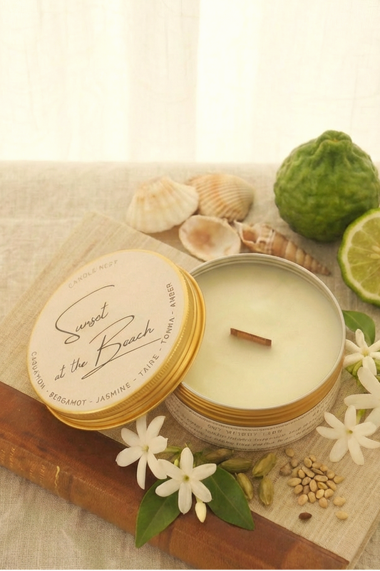 Ett doftljus i en rund guldfärgad burk av metall står på en bok. Boken är av linnetyg med en rygg i läder. Ljusets lock står lutad på ljuset på på det finns ljusets namn skrivet med snirklig text. Mitt i ljuset syns en veke av trä. Runt ljuset syns snäckor, vita små blommor, bergamott och kardemumma kärnor. 