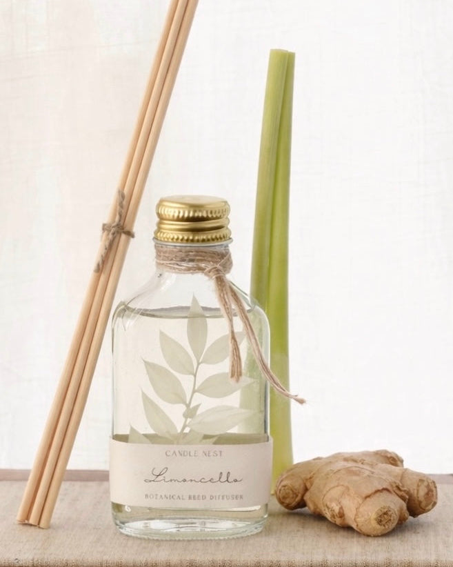 En genomskinlig flaska med doftpinnar står på ett bord. I flaskan syns en gren med vita blad. Flaskan har en skruvkork av guld och pinnarna (reeds)står lutade mot flaskan. Citrongräs står lutat mot flaskan och bredvid flaskan ligger en bit ingefära.