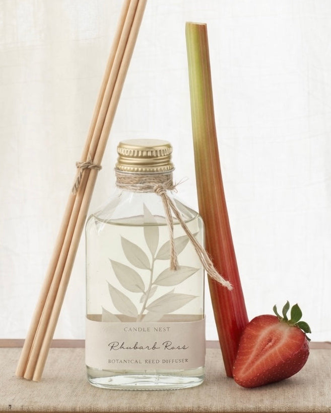 En genomskinlig flaska med doftpinnar står på ett bord. I flaskan syns en gren med vita blad. Flaskan har en skruvkork av guld och pinnarna (reeds)står lutade mot flaskan. En stjälk rabarber står lutad mot flaskan och bredvid flaskan ligger en jordgubbe