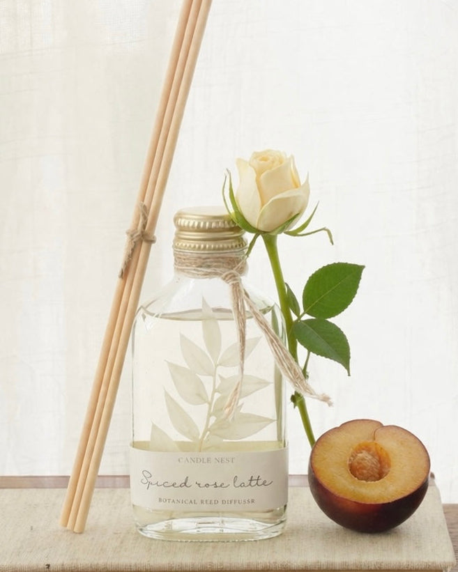 En genomskinlig flaska med doftpinnar står på ett bord. I flaskan syns en gren med vita blad. Flaskan har en skruvkork av guld och pinnarna (reeds)står lutade mot flaskan. En vi ros står lutad mot flaskan och bredvid rosen ligger ett plommon.