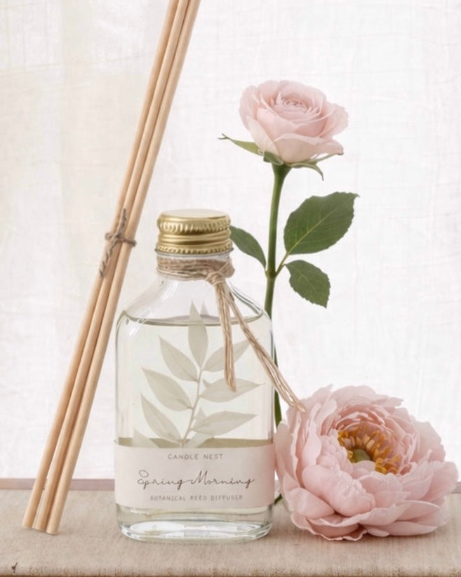 En genomskinlig flaska med doftpinnar står på ett bord. I flaskan syns en gren med vita blad. Flaskan har en skruvkork av guld och pinnarna (reeds)står lutade mot flaskan. En rosa ros står lutad mot flaskan och bredvid flaskan ligger en pion