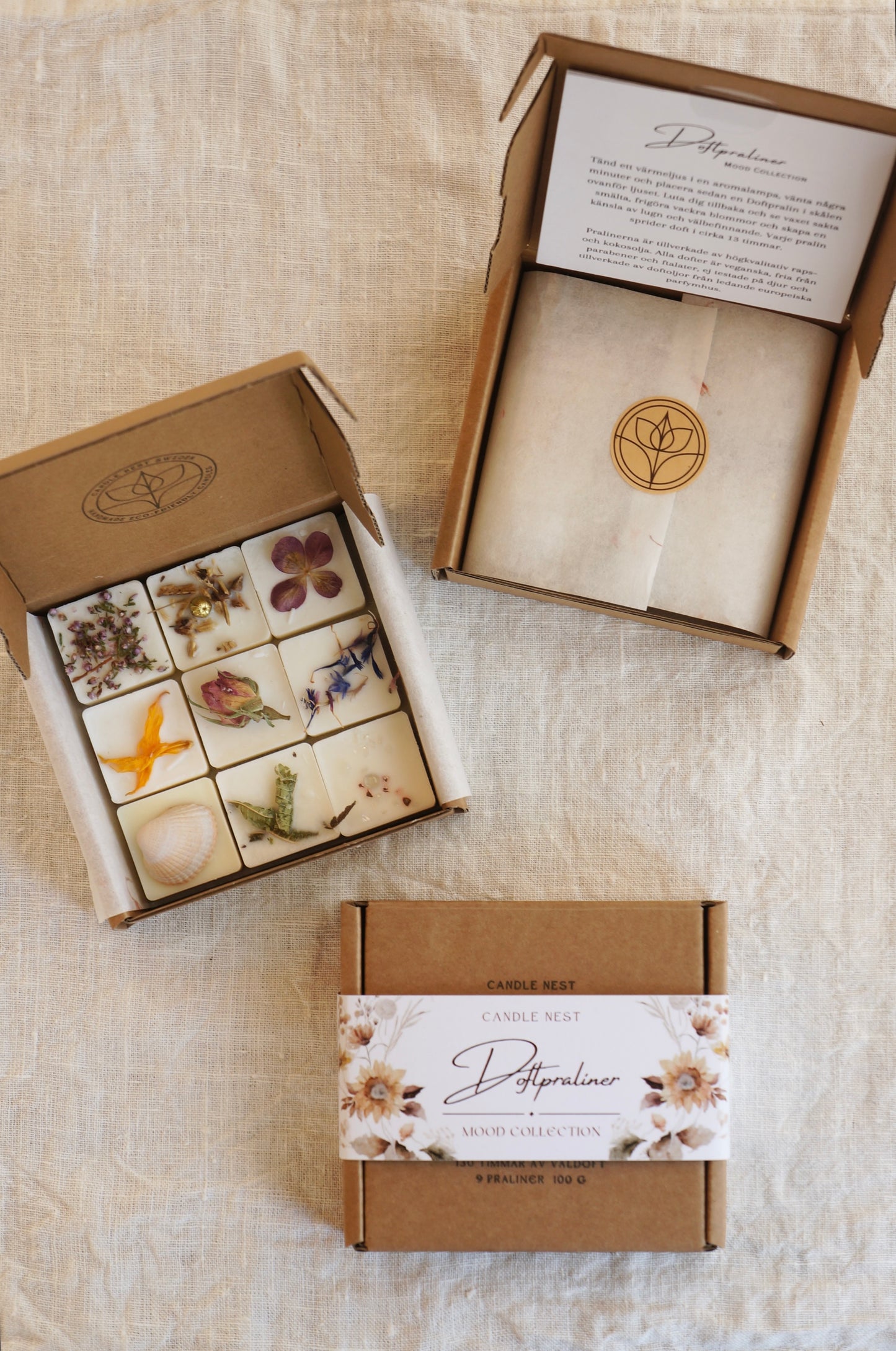 Tre askar med Doftpraliner ligger på ett bord. En är öppen och blottar Doftpralinerna som är dekorerade med torkade blommor. En ask är stängd och en är öppen där man ser pappret med instruktionens som sitter fast på insidan av locket. Pralinerna täcks av ett papper med riktiga blommor infattade i pappret. 