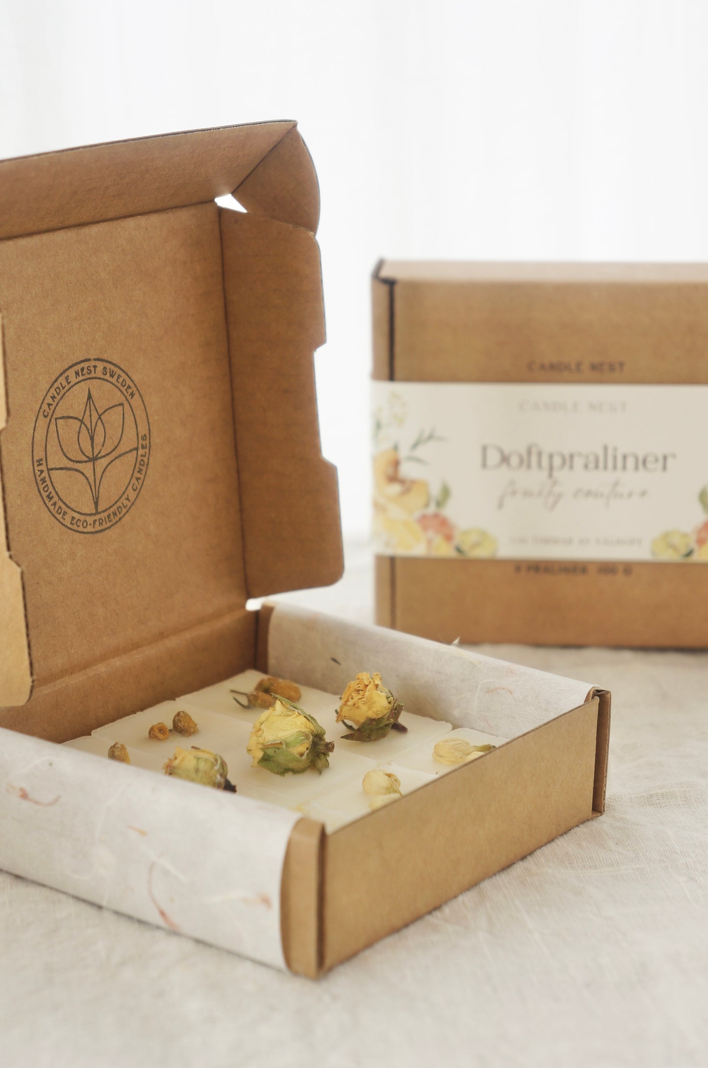 En naturfärgad ask med doftpraliner står på ett bord. Runt asken finns ett papper med texten "Doftpraliner Fruity couture" på. Pappret är dekorerat med frukter, målade i vattenfärg. I förgrunden syns en ask med doftpraliner dekorerade med torkade blommor  