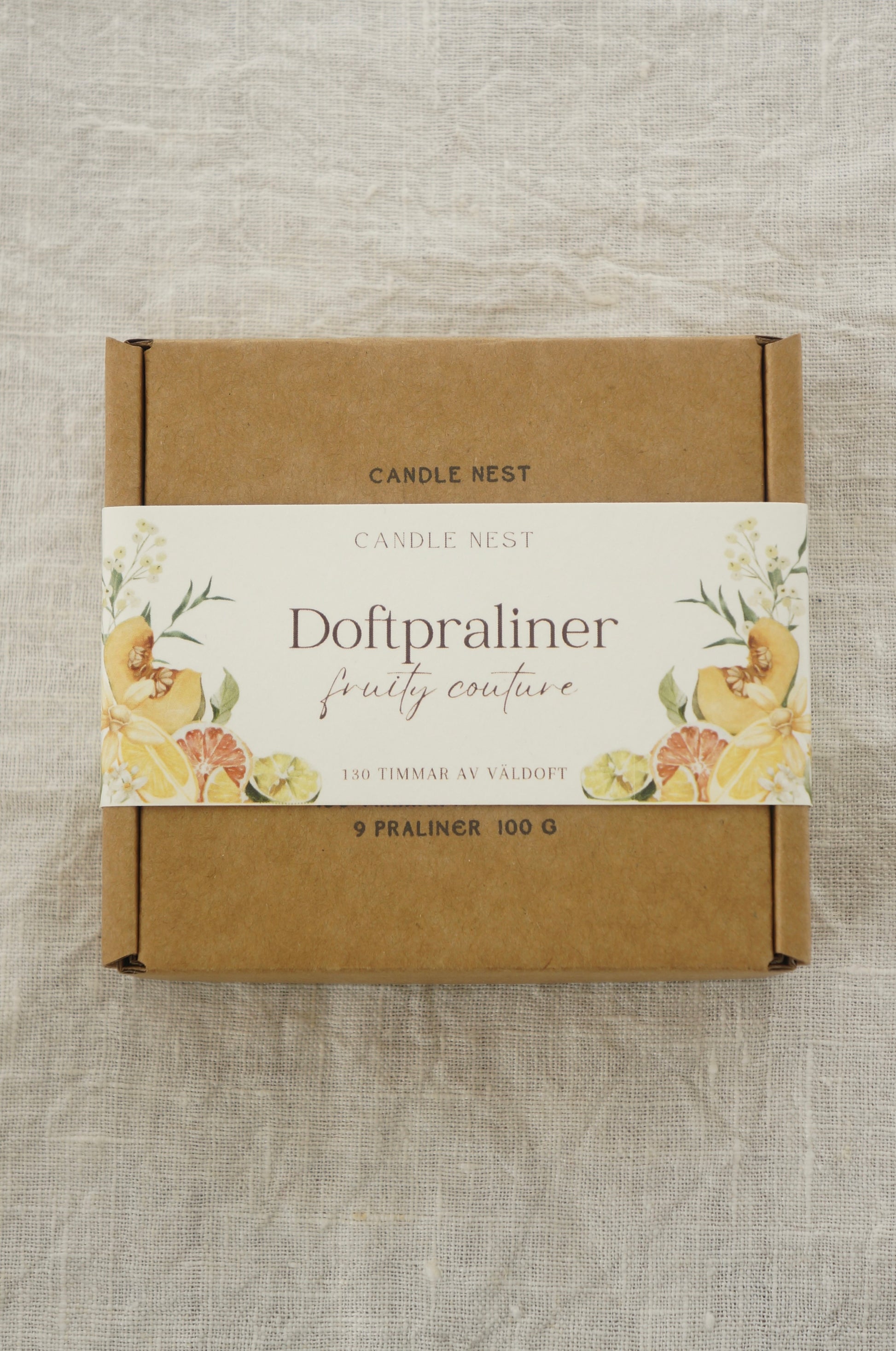 En brun ask innehållandes doftvax  ligger på en duk av linnetyg. På asken finns texten "Doftpraliner- Fruity couture".  