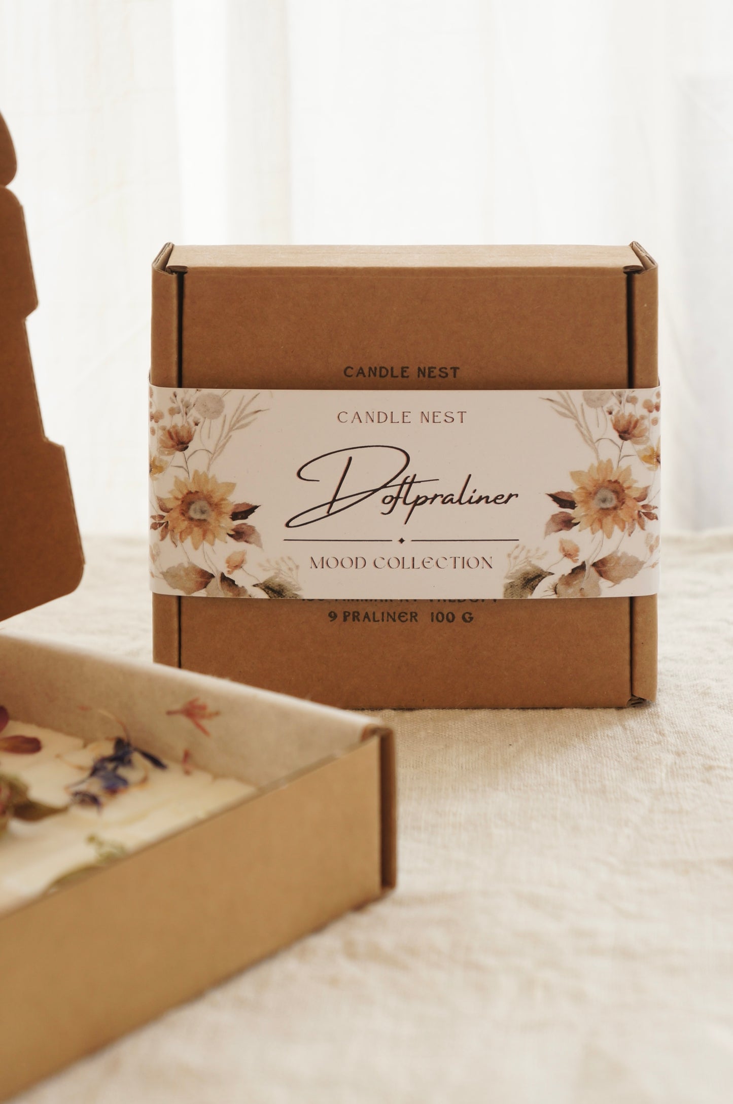 En naturfärgad ask med doftpraliner står på ett bord. Runt asken finns ett papper med texten doftpraliner Mood Collection på. Pappret är dekorerat med blommor, målade i vattenfärg i gult och beiget och brunt. I förgrunden syns en ask med doftpraliner dekorerade med torkade blommor  