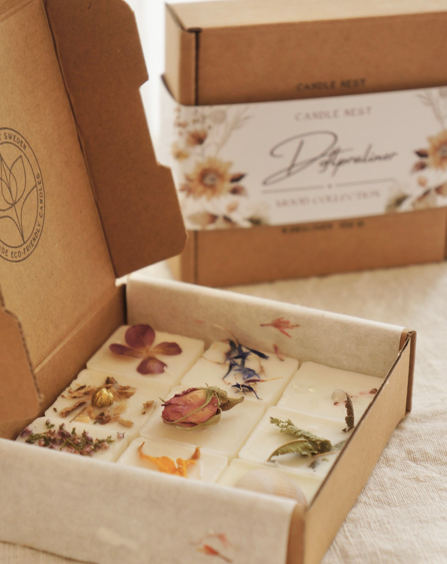 En naturfärgad ask med doftpraliner står på ett bord. Runt asken finns ett papper med texten doftpraliner Mood Collection på. Pappret är dekorerat med blommor, målade i vattenfärg i gult och beiget och brunt. I förgrunden syns en öppen ask med doftpraliner dekorerade med torkade blommor.   
