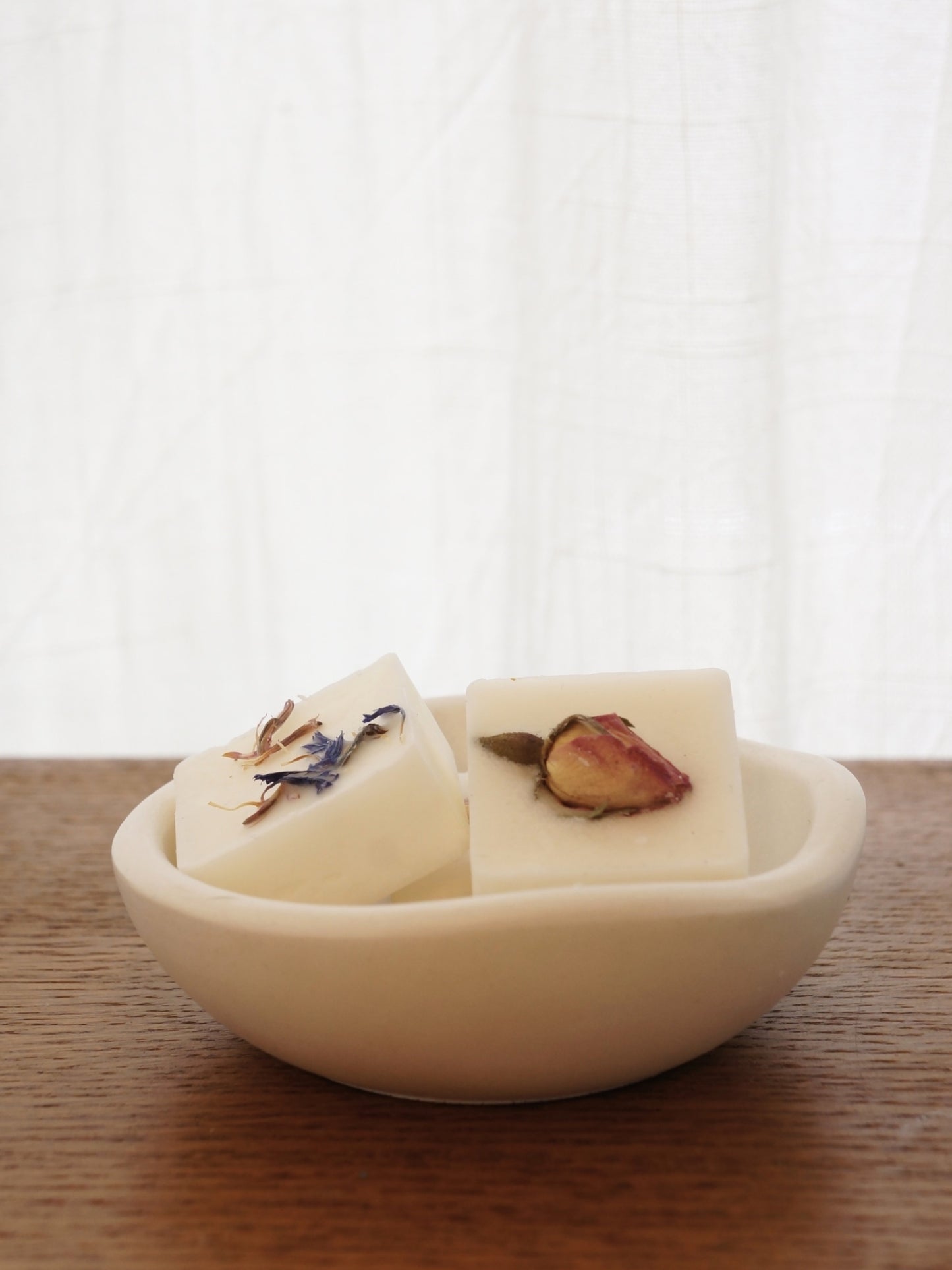 En liten vit skål i gips med vågformad kant står på ett mörkt bord av trä. I skålen ligger två doftpraliner dekorerade med torkade blommor, ros och blåklint. 