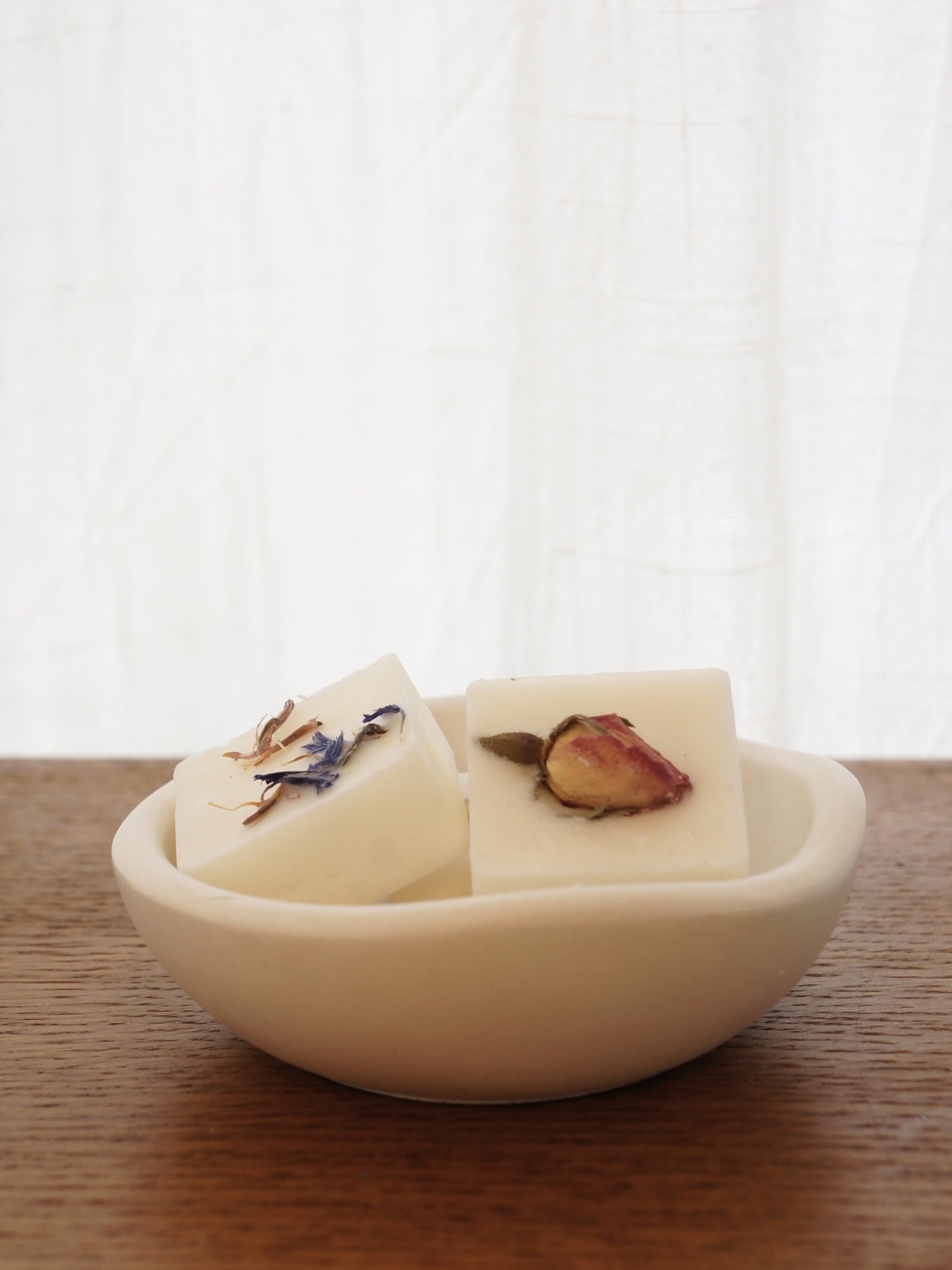 En liten vit skål i gips med vågformad kant står på ett mörkt bord av trä. I skålen ligger två doftpraliner dekorerade med torkade blommor, ros och blåklint. 