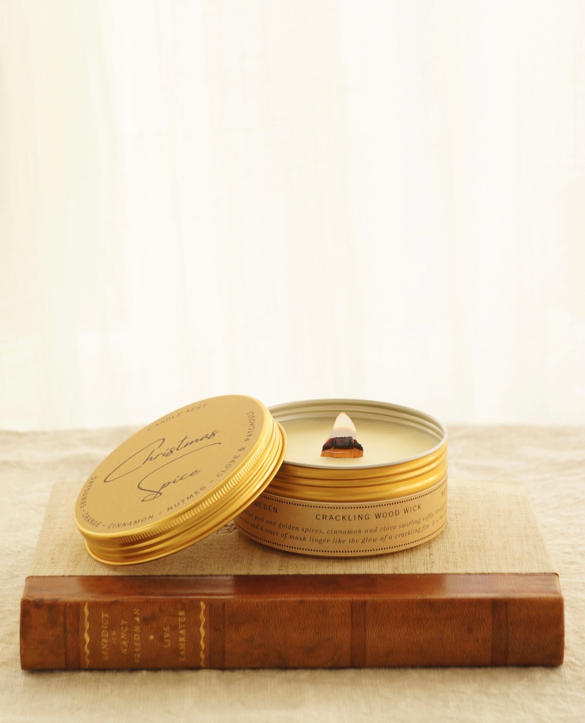 Ett doftljus i guldig aluminiumburk med en veke av trä står på en äldre bok med rygg av läder. Boken ligger på ett bord, täckt med en vit duk av linnetyg