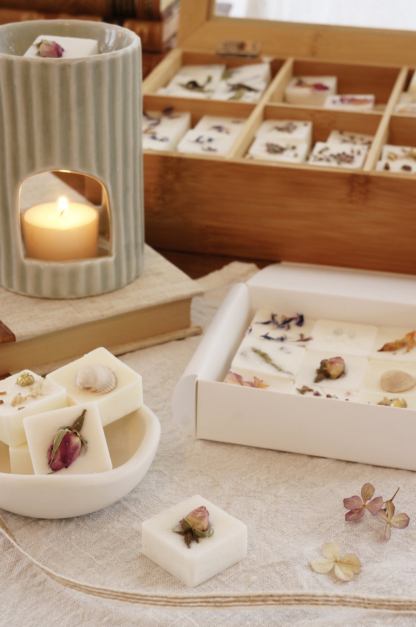 En aromalampa, en skål med doftpraliner står på ett en beige duk av linnetyg. I bakgrunden syns en telåda i trä innehållandes doftpraliner. Ljuset är varmt, mjukt och inbjudande. 