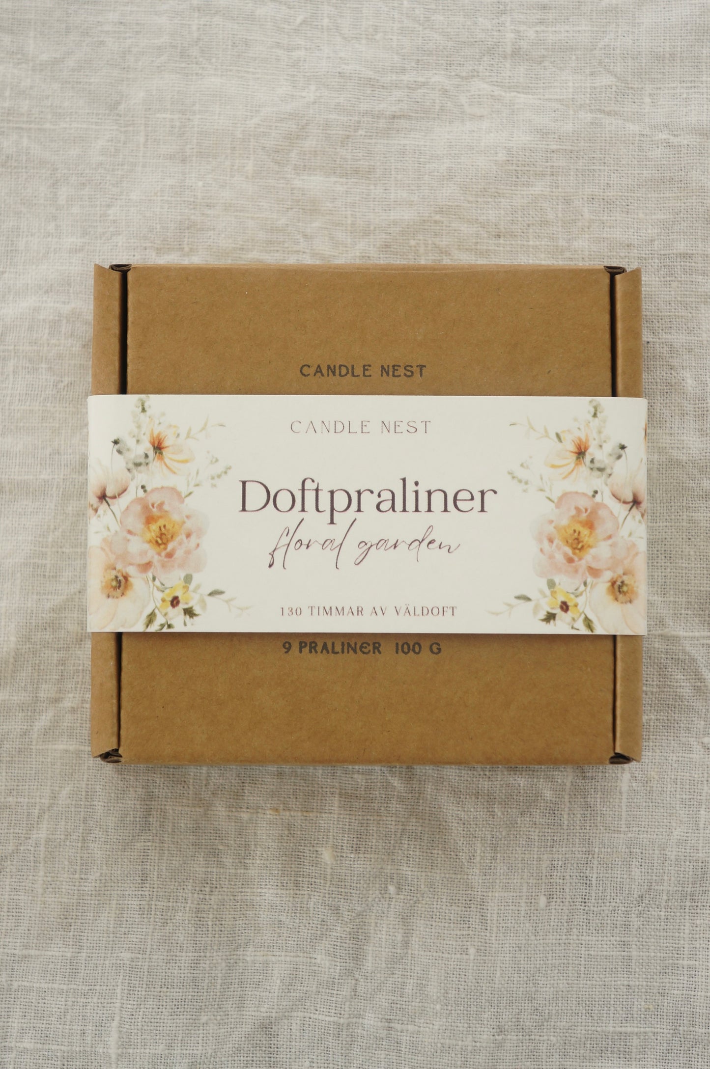 En brun ask innehållandes doftvax  ligger på en duk av linnetyg. På asken finns texten "Doftpraliner- Floral garden".  