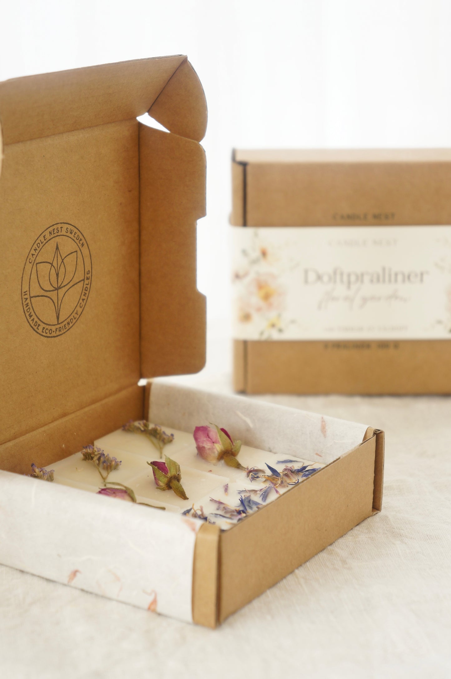 En naturfärgad ask med doftpraliner står på ett bord. Runt asken finns ett papper med texten "Doftpraliner Floral garden" på. Pappret är dekorerat med blommor, målade i vattenfärg. I förgrunden syns en ask med doftpraliner dekorerade med torkade blommor  
