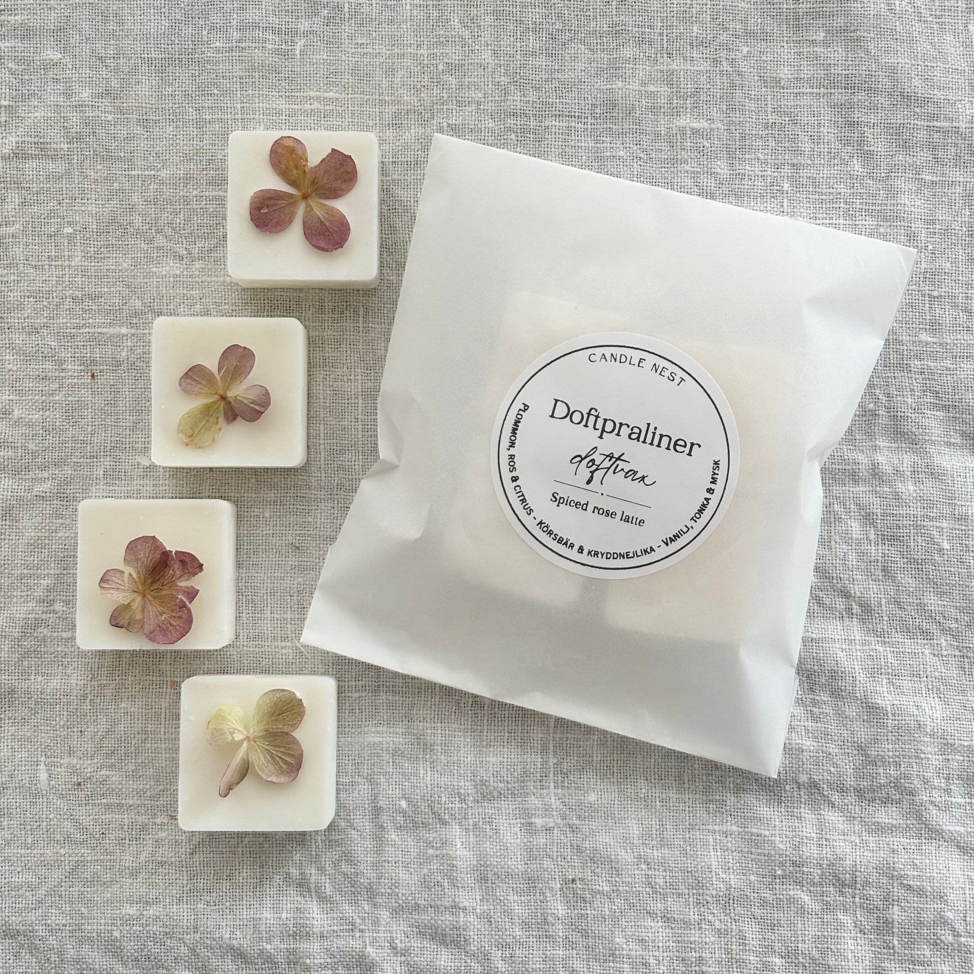 En vit semi transparent påse innehållandes doftvax (doftpraliner) ligger på en linneduk. Runt påsen finns fyra vita doftpraliner utplacerade. Doftvaxen är dekorerade med ekologiskt odlad hortensia.