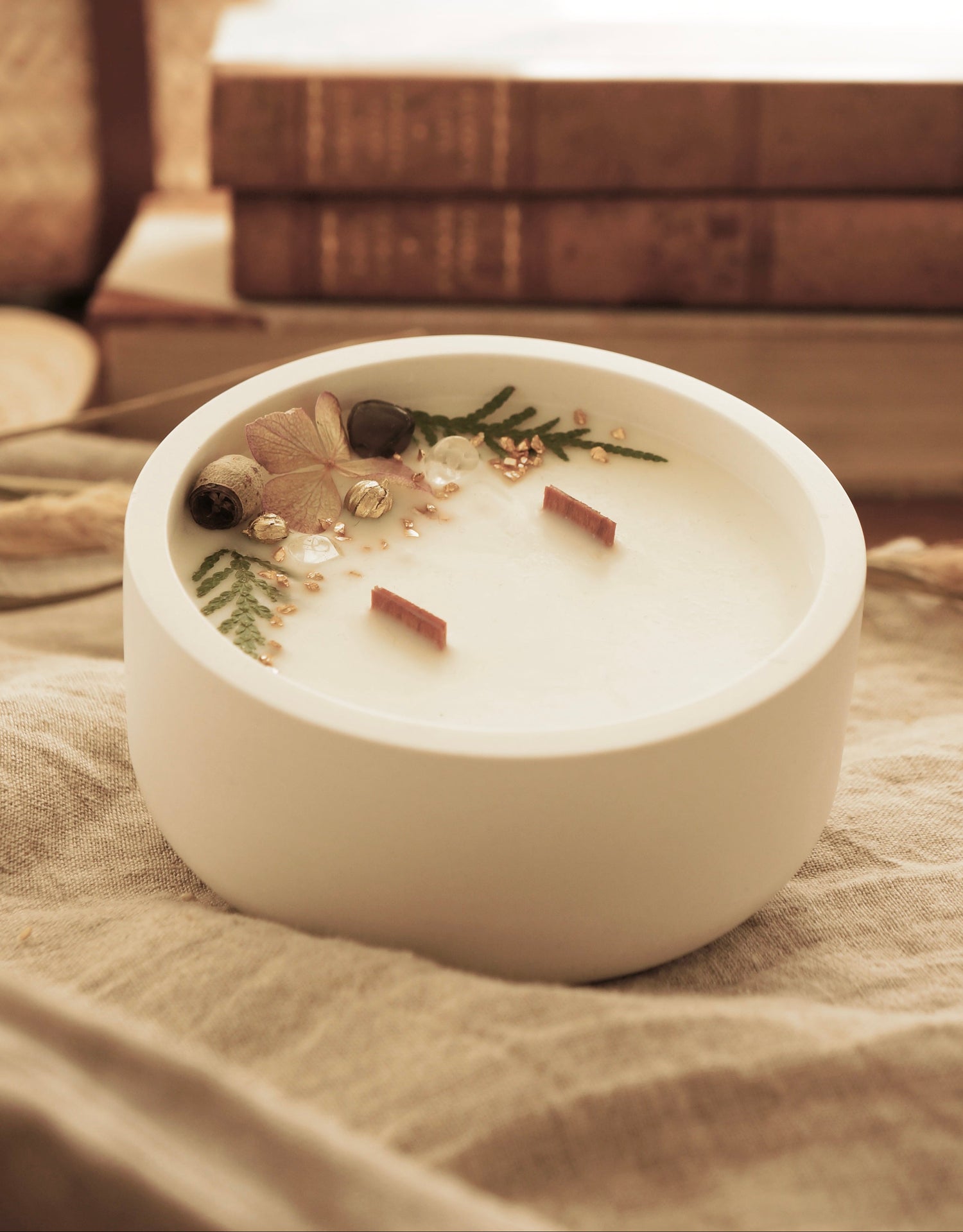 Ett doftljus står på en lätt skrynklad linneduk. I bakgrunden syns äldre böcker. Ljuset är mjukt. Doftljuset är dekorerat med orkade växter. 