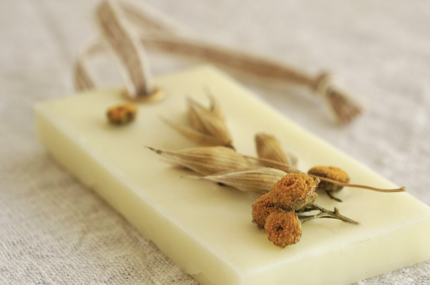 Dofthänge av raps- och koksvax i doften "Farmers market" som ligger på en ljus beige linneduk. Dekroerade med torkad havre och renfana. Närbild tagen snett från sidan.