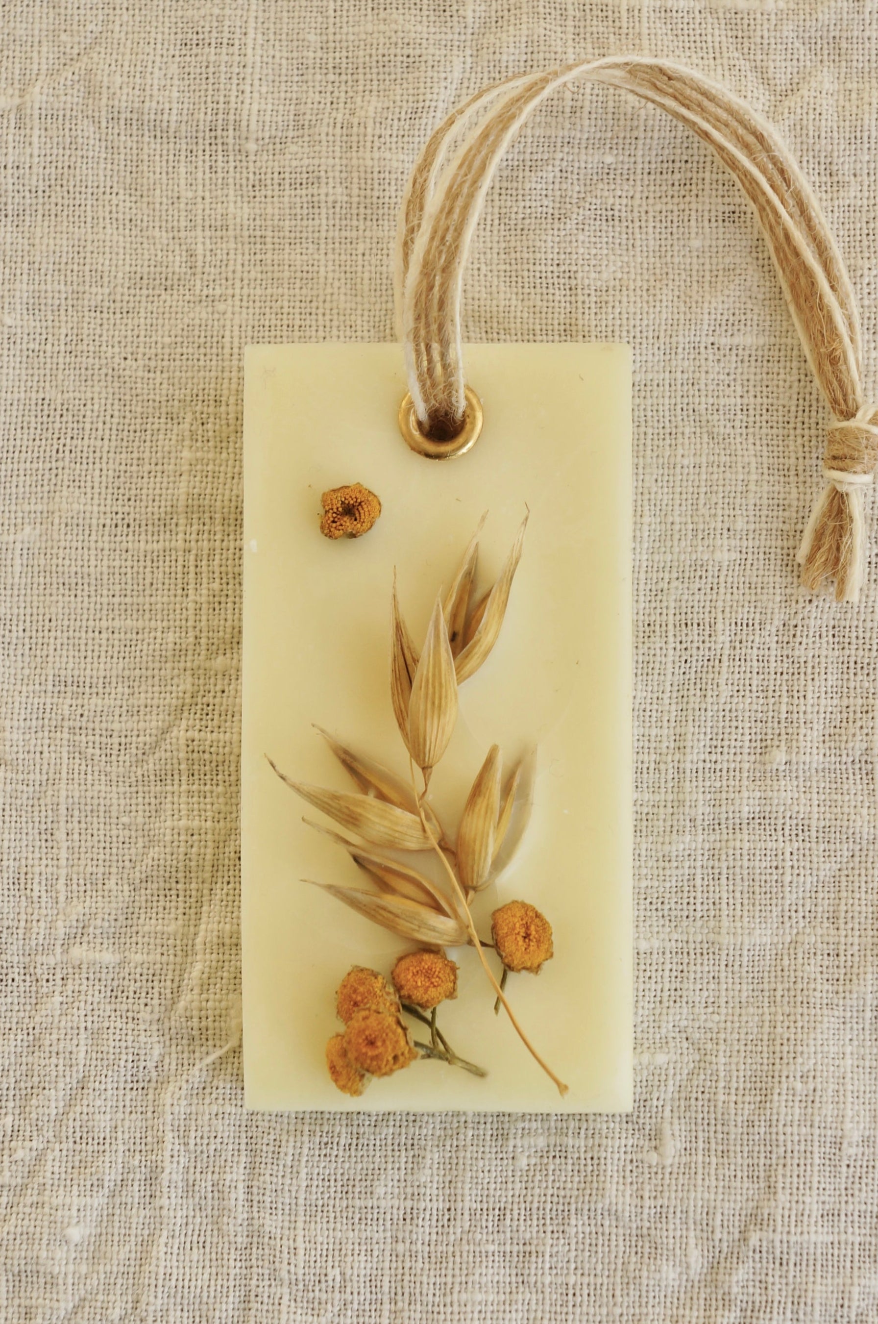 Dofthänge av raps- och koksvax i doften "Farmers market" som ligger på en ljus beige linneduk. Dekorerad med torkad havre och renfana. Bild tagen ovanifrån.