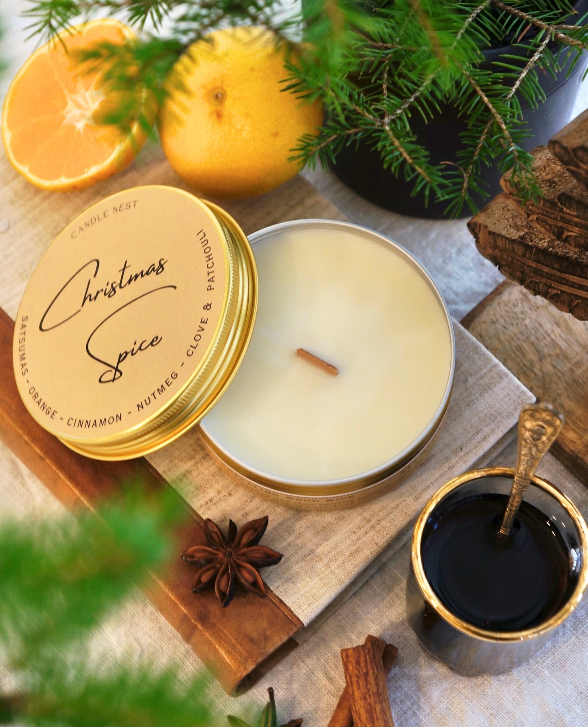 Ett doftljus i en guldig aluminiumburk står på en bok av linnetyg Med läderrygg. Ljuset är omgivet av Clementiner, Kanel, stjärnanis och granris. Bredvid ljuset står en glögg kopp i glas med en liten sked i.