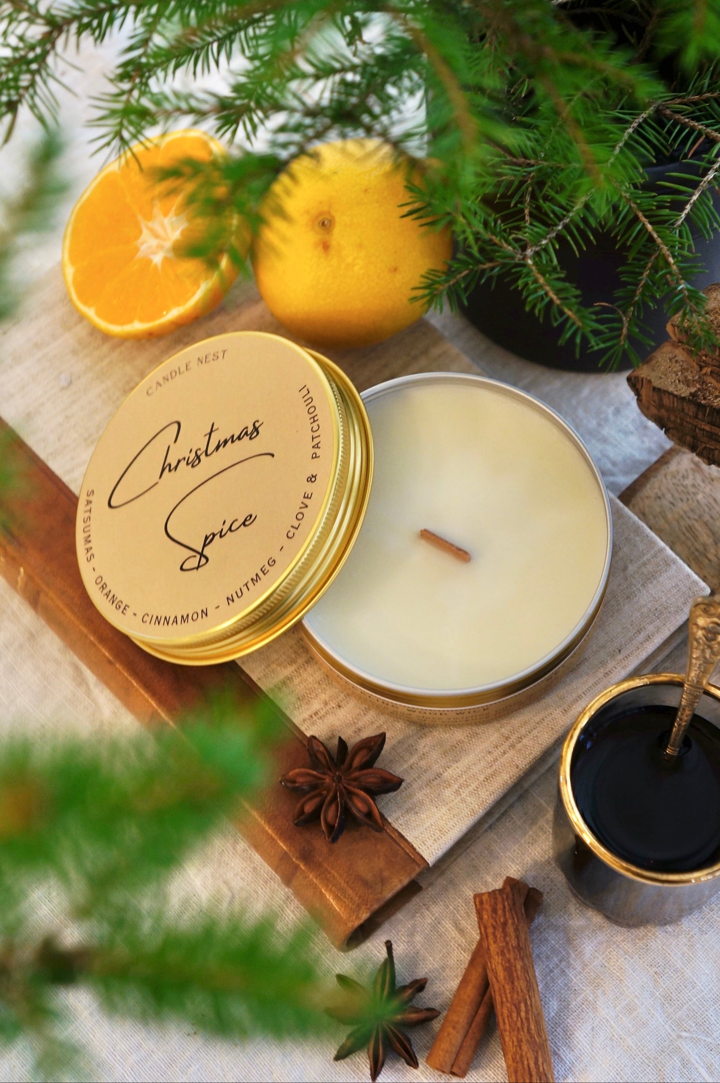 Ett doftljus i en guldig aluminiumburk står på en bok av linnetyg Med läderrygg. Ljuset är omgivet av Clementiner, Kanel, stjärnanis och granris. Bredvid ljuset står en glögg kopp i glas med en liten sked i.