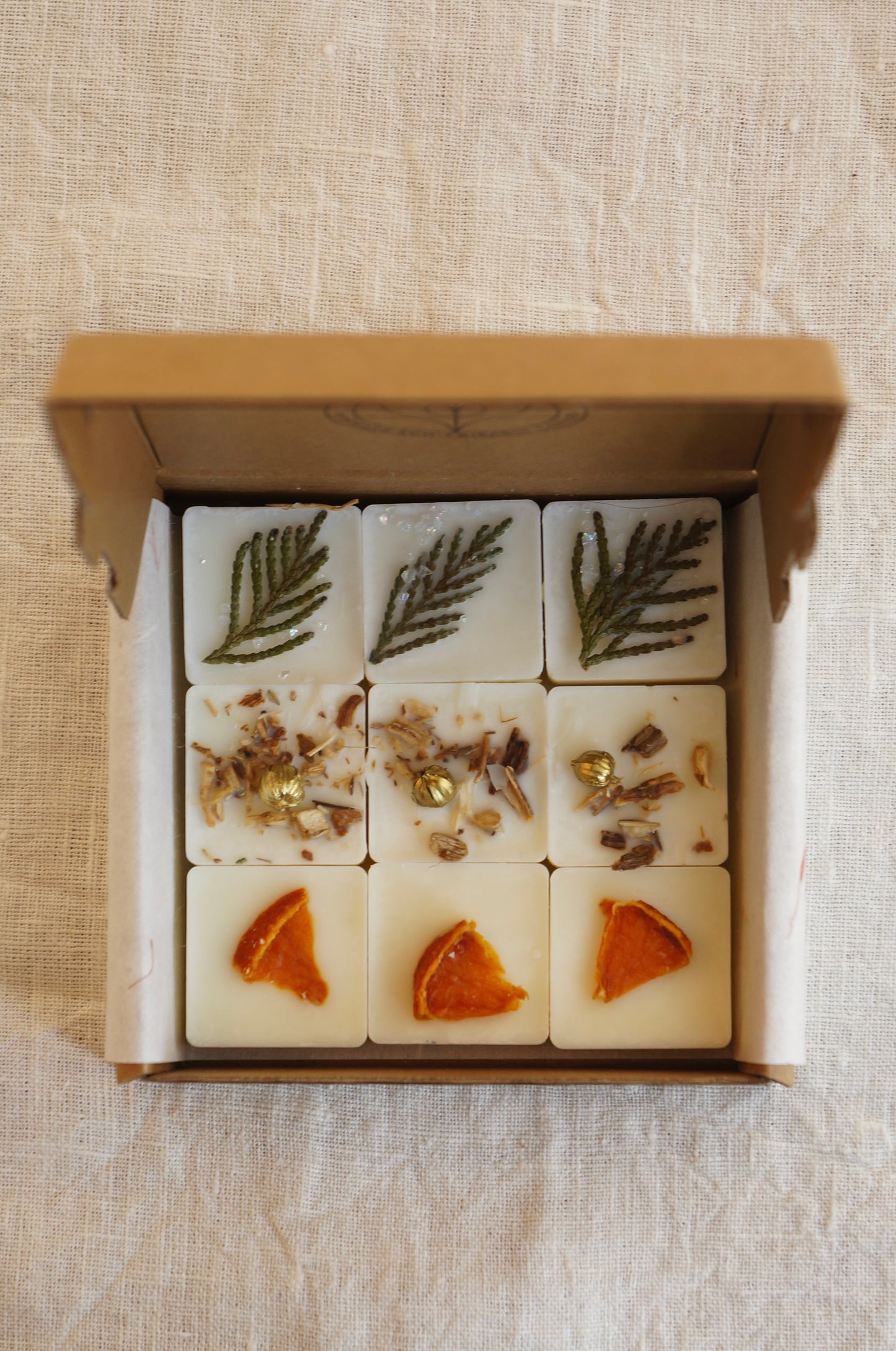 En ask i naturfärgad papp innehållandes 9 st doftvax (doftpraliner) står på ett bord. Doftpralinerna av raps och kokosvax är dekorerade med torkade bitar av clementin och små bitar av thuja. 