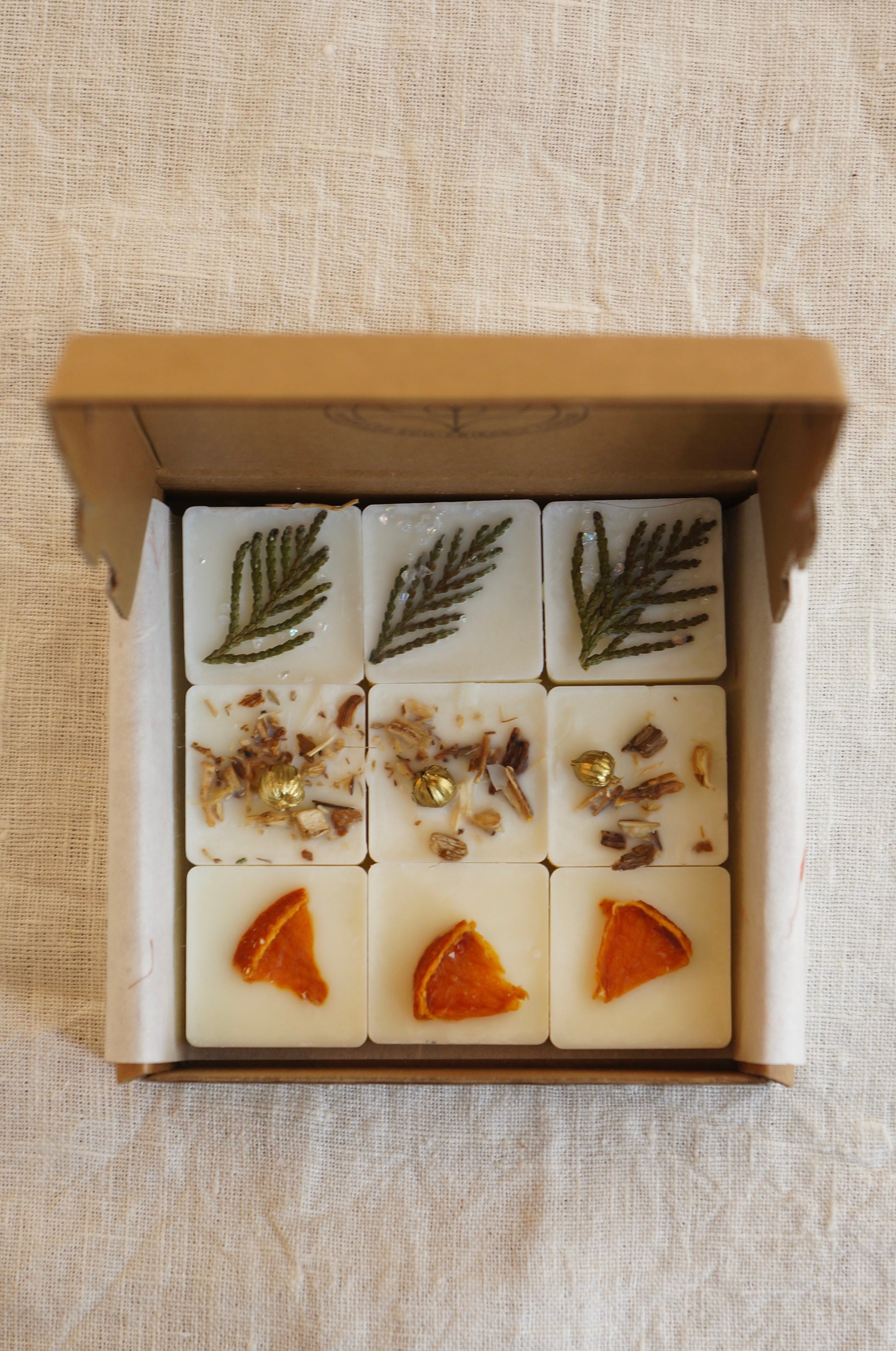 En ask i naturfärgad papp innehållandes 9 st doftvax (doftpraliner) står på ett bord. Doftpralinerna av raps och kokosvax är dekorerade med torkade bitar av clementin och små bitar av thuja. 