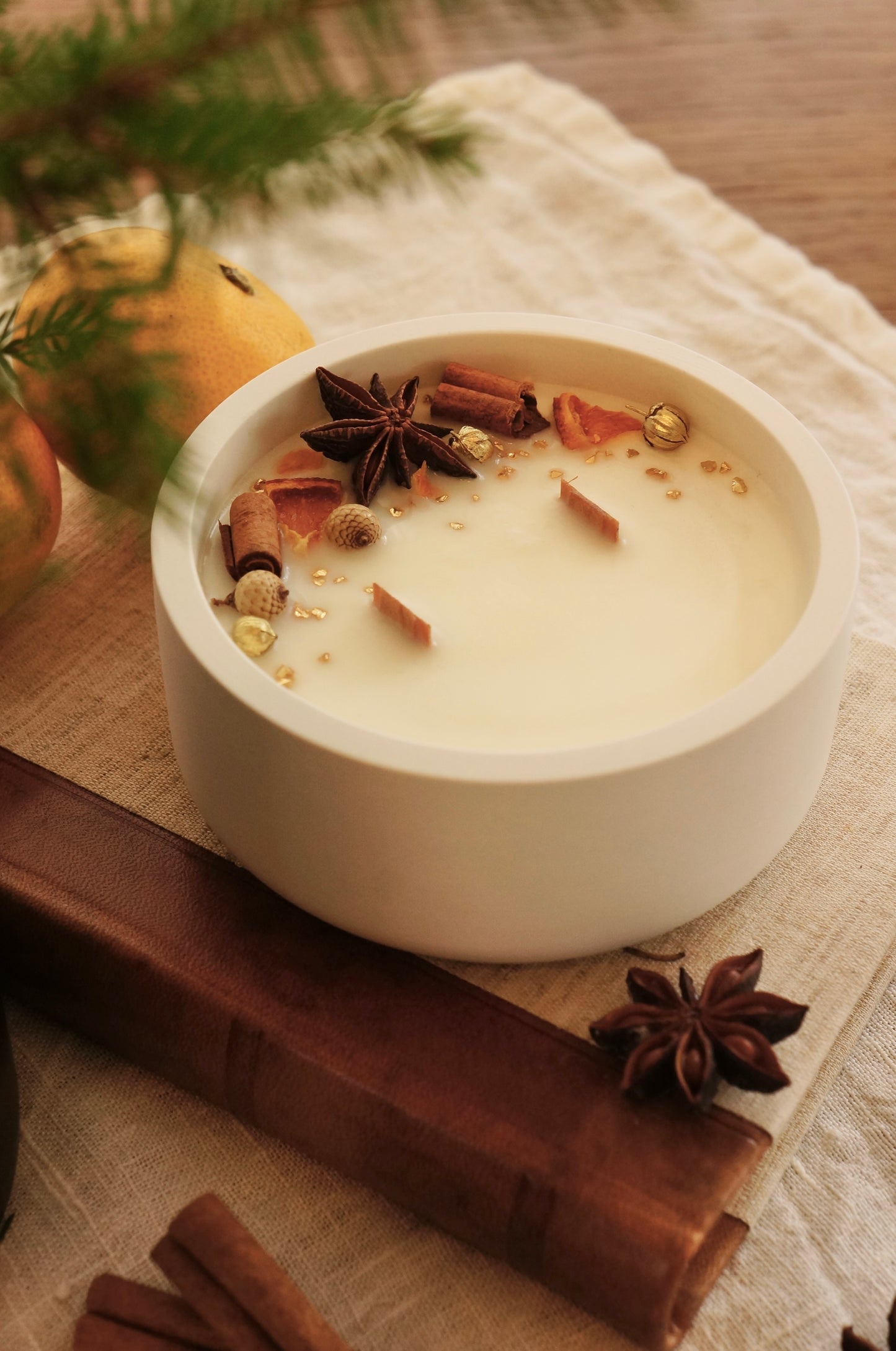 Doftljus i en vit gispskål står på en äldre bok med läderrygg. Ljusets ovansida är dekorerat med små bitar av clementin, kanlestänger och guldfärgade linfrökapsalar. i bakgrunden syns en grankvist och clementiner.