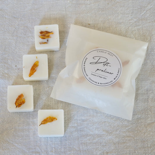 Fyra vita doftpraliner med doften "Dreams of Napa Valley" dekorerade med Torkade kronblad av solros från vår trädgård. Doftvaxen är arrangerade på ett vitt linnetyg bredvid en transparent förpackningspåse från Candle Nest