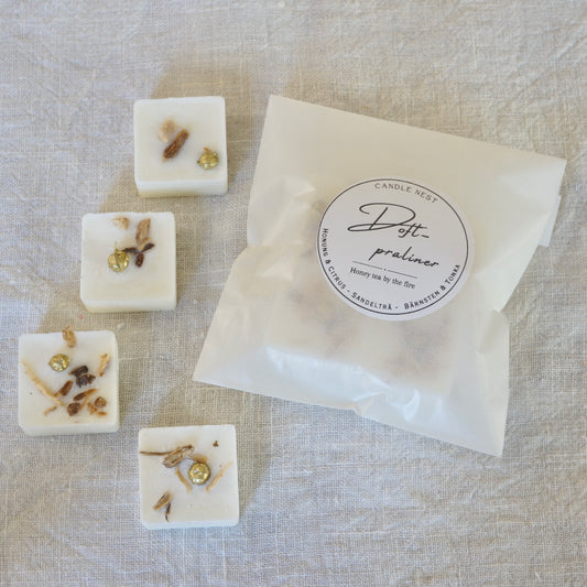 Fyra vita doftpraliner med doften "Honey tea by the Fire" dekorerade med Ekologiskt odlad cikoriarot och torkade linfrökapslar. Doftvaxen är arrangerade på ett vitt linnetyg bredvid en transparent förpackningspåse från Candle Nest