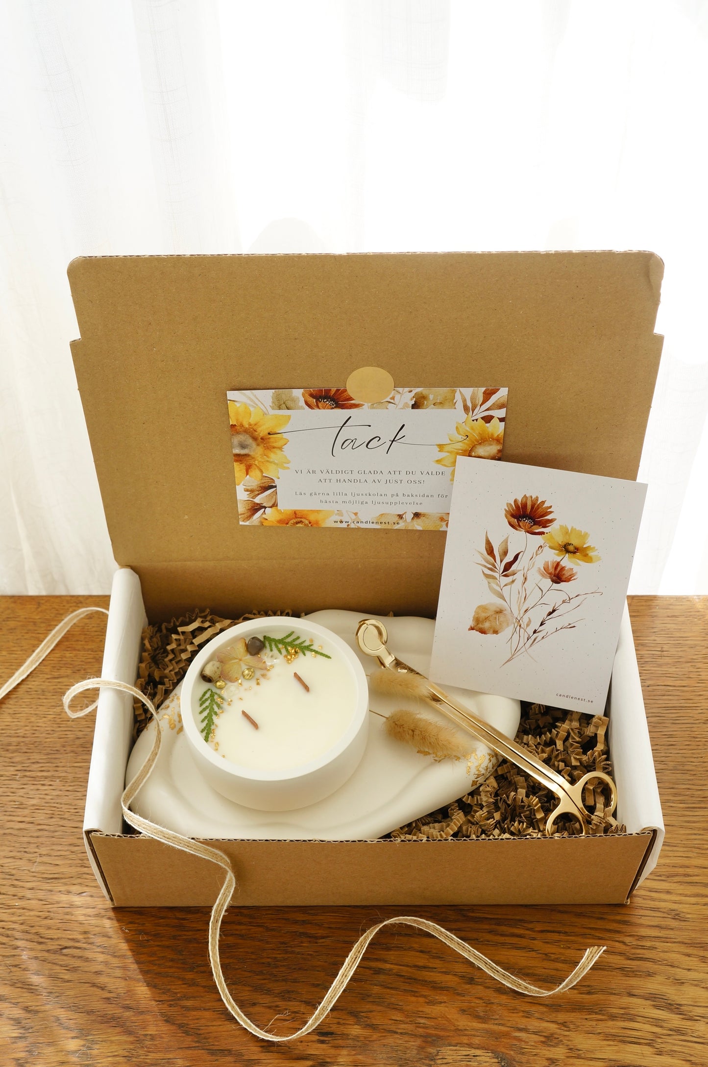 Presentbox med Botaniskt doftljus (Honey tea by the fire), ljusfat med gyllen dekor och en vektång. Produkterna ligger i en låda av naturfärgd papp. 