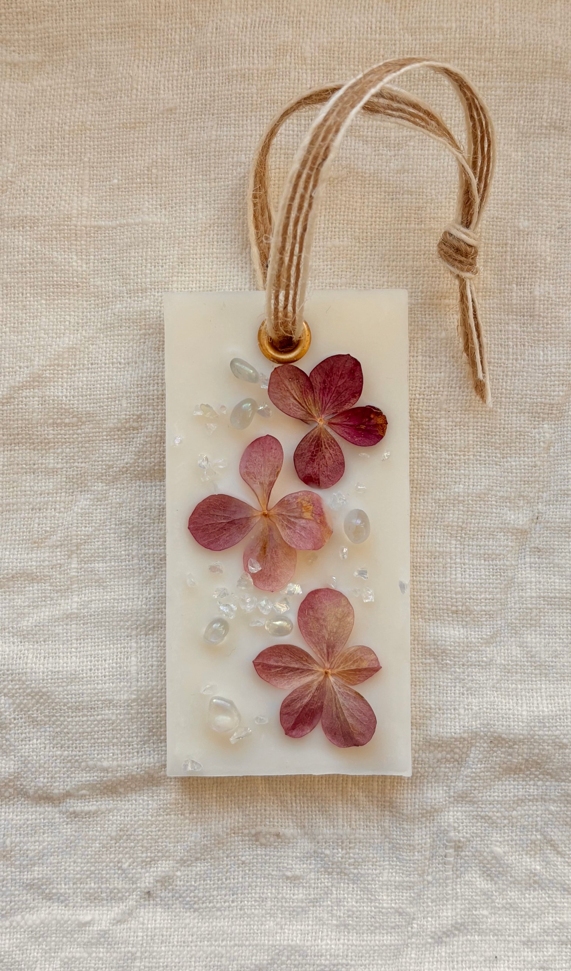 Rektangulärt Dofthänge av vax ligger på en vit linneduk. Dofthängen är dekorerat med torkad hortensia och glaspärlor 