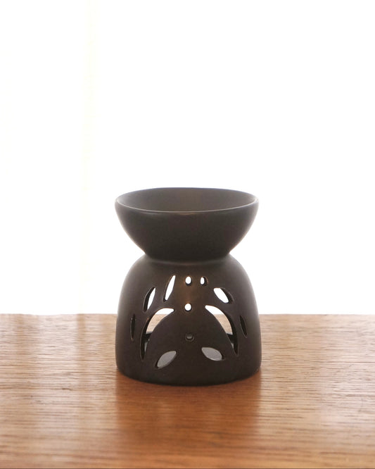 En liten svart aromalampa i oval form står på ett bord av trä. På lampans framsida syns ett dekorativt utskuret mönster. 