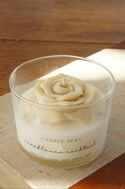 Candle nest doftljus, casablanca cocktail. Transparent glas, en botten av vitt vax och en ljusbeige ros på toppen. veka av bomull i mitten av rosen. Ljuset står på en äldre bok som ligger på ett bord av trä. Bild tagen snett uppifrån