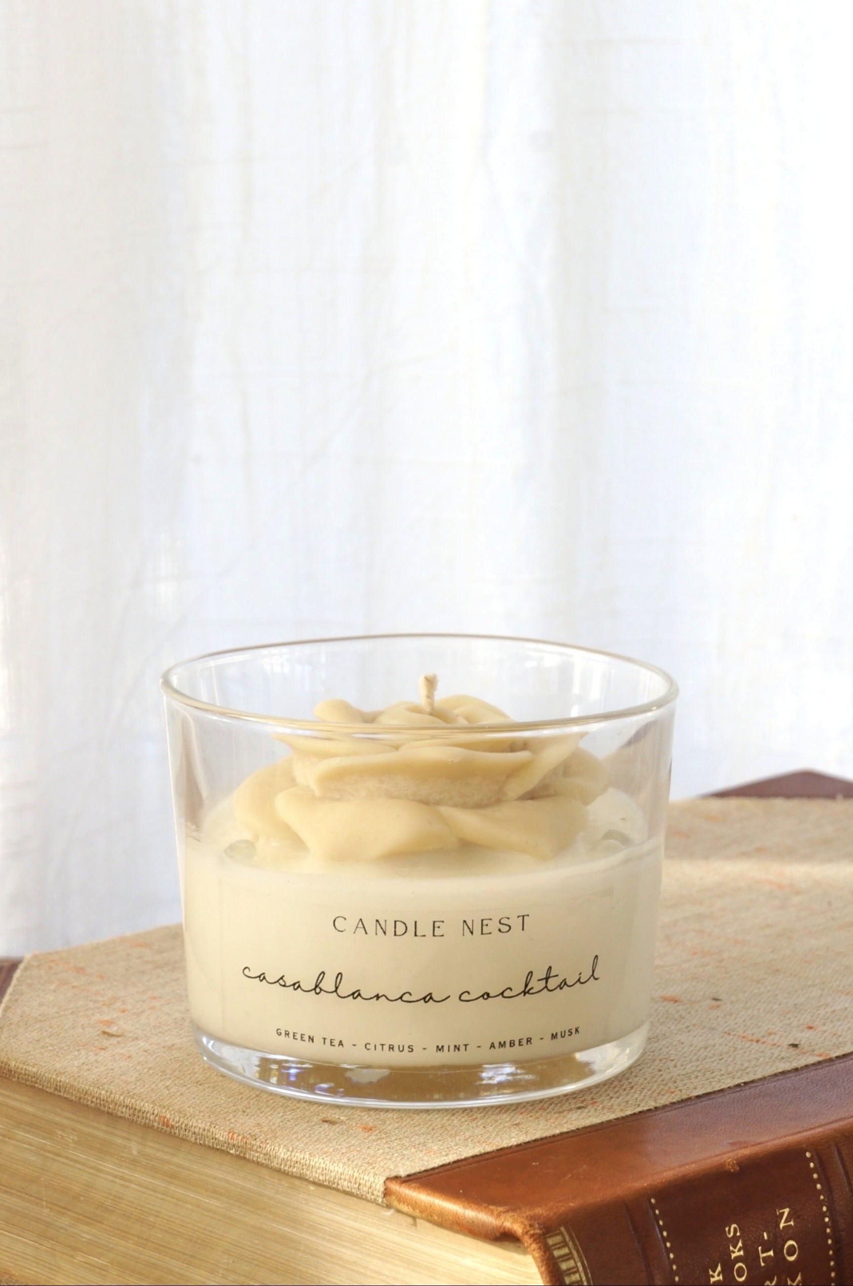 Candle nest doftljus, casablanca cocktail. Transparent glas, en botten av vitt vax och en ljusbeige ros på toppen. veka av bomull i mitten av rosen. Ljuset står på en äldre bok som ligger på ett bord av trä. Bild tagen från sidan.