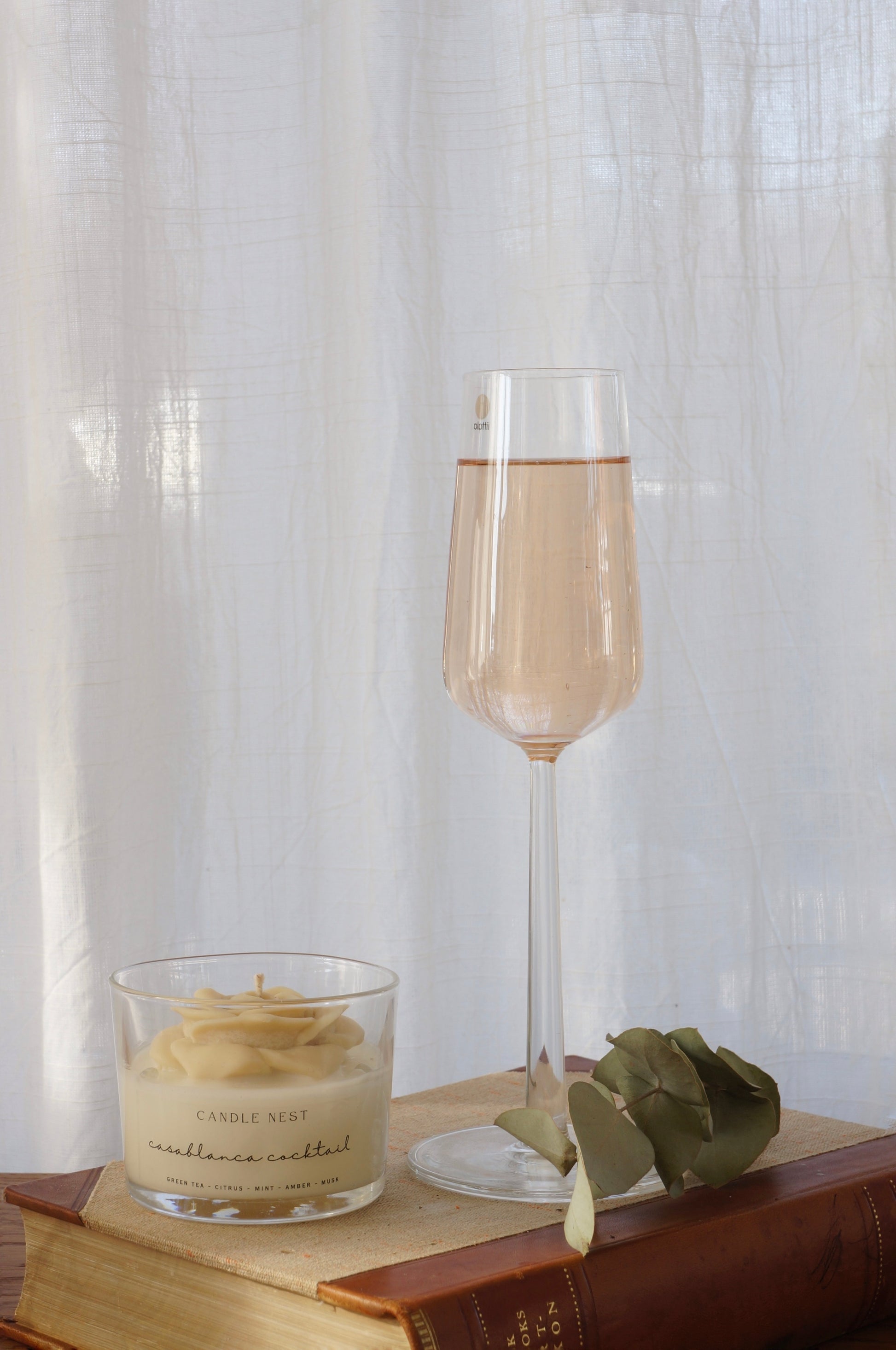 Candle nest doftljus, casablanca cocktail. Transparent glas, en botten av vitt vax och en ljusbeige ros på toppen. veka av bomull i mitten av rosen. Ljuset står på en äldre bok och bredvid står en ljusrosa transparent drink i ett högt glas. En kvist av torkad eukalyptus ligger bredvid glaset. 