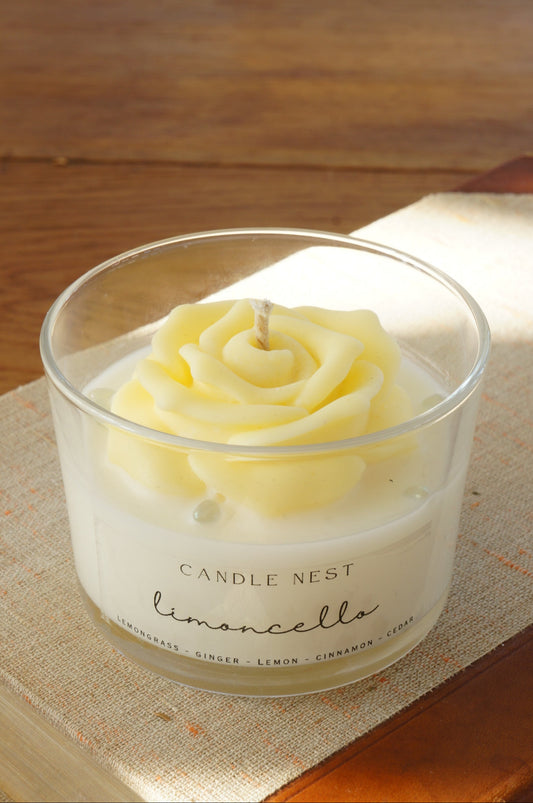 Candle nest doftljus, Limoncello. Transparent glas, en botten av vitt vax och en ljusgul ros av vax på toppen. veke av bomull i mitten av rosen. Ljuset står på en äldre bok. Bild tagen snett uppifrån.