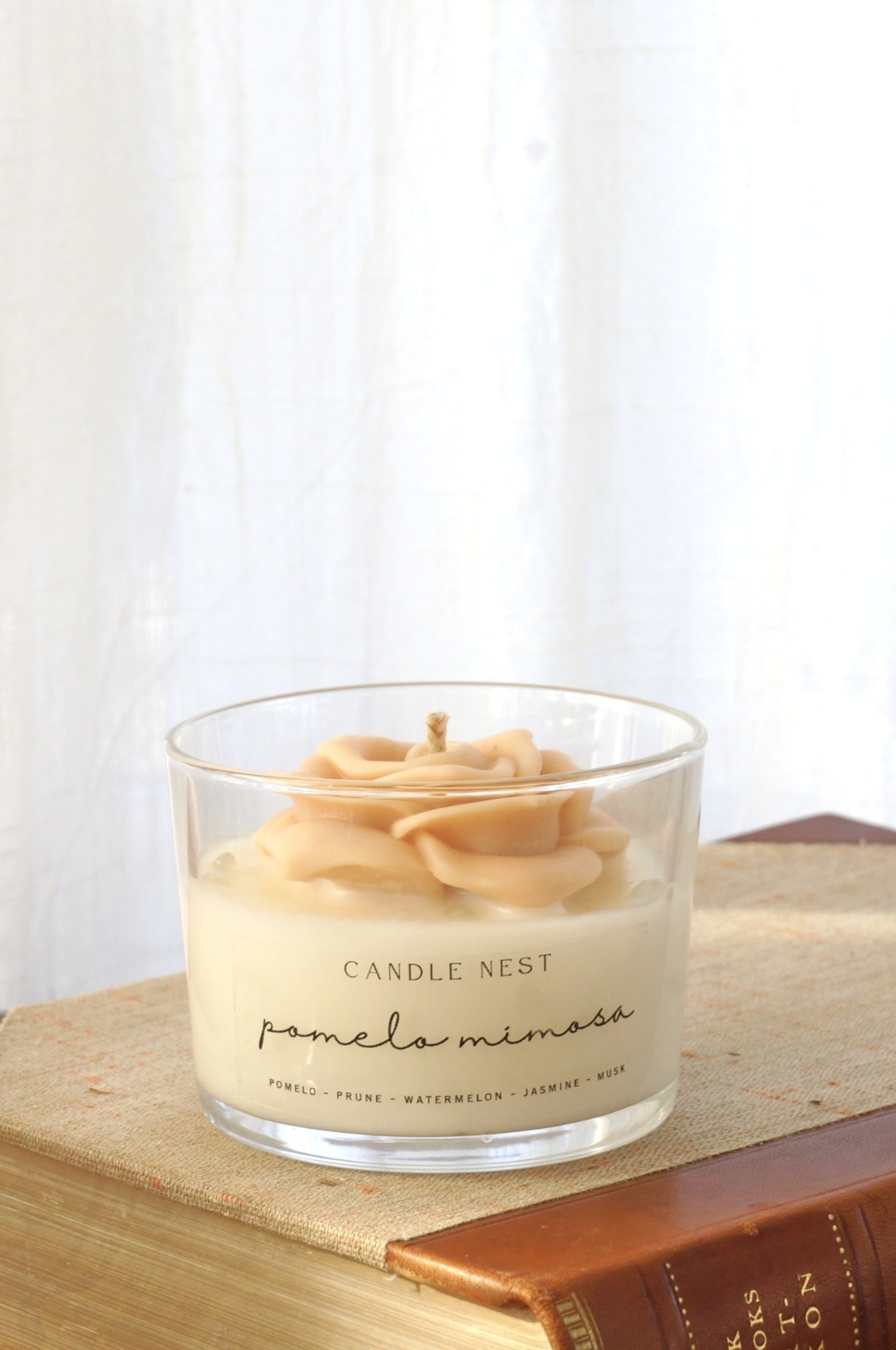 Candle nest doftljus, Pomelo mimosa. Transparent glas, en botten av vitt vax och en ljusrosa ros av vax på toppen. veke av bomull i mitten av rosen. Ljuset står på en äldre bok. Bild tagen framifrån.
