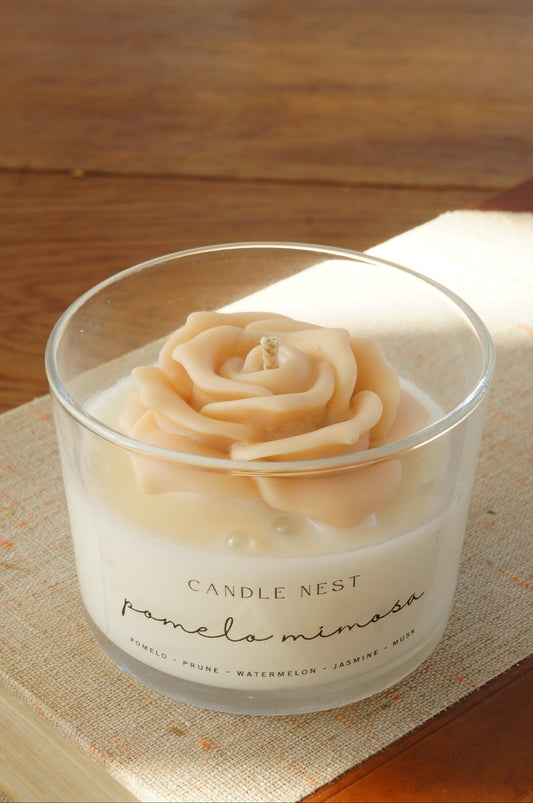 Candle nest doftljus, Pomelo mimosa. Transparent glas, en botten av vitt vax och en ljusrosa ros av vax på toppen. veke av bomull i mitten av rosen. Ljuset står på en äldre bok. Bild tagen snett uppifrån.