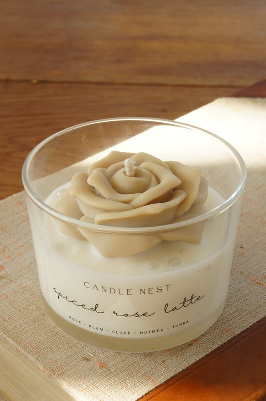 Candle nest doftljus, Spiced rose latte. Transparent glas, en botten va vitt vax och en beige ros på toppen. veka av bomull i mitten av rosen. Ljuset står på en äldre bok som ligger på ett bord av trä
