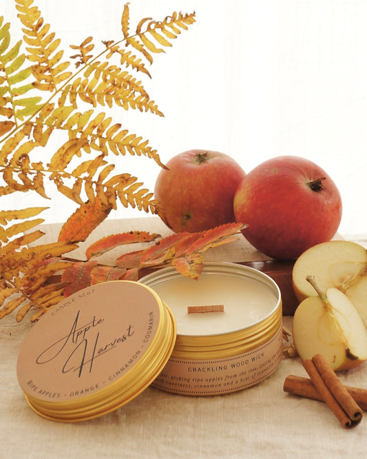Candle nest doftljus "Apple Harvest" i en rund guldig burk av metall står på ett bord omgivet av äpplen,, kanel och höstliga löv och kvistar. Locket är lutat mot sidan av burken och vax och veke av trä är exponerat. 