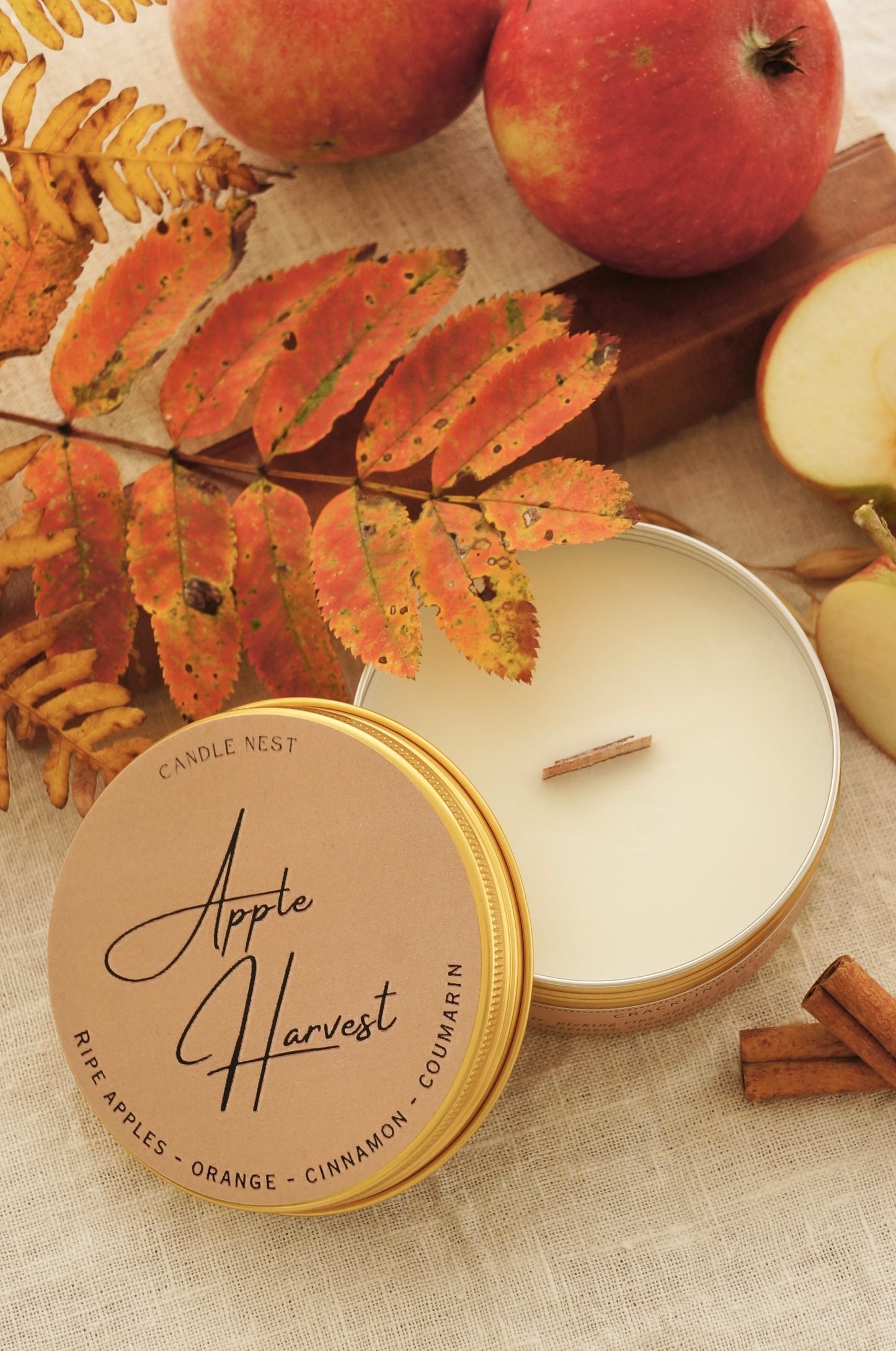 Candle nest doftljus "Apple Harvest" i en rund guldig burk av metall står på ett bord omgivet av äpplen,, kanel och höstliga löv och kvistar. Locket är lutat mot sidan av burken och vax och veke av trä är exponerat. 